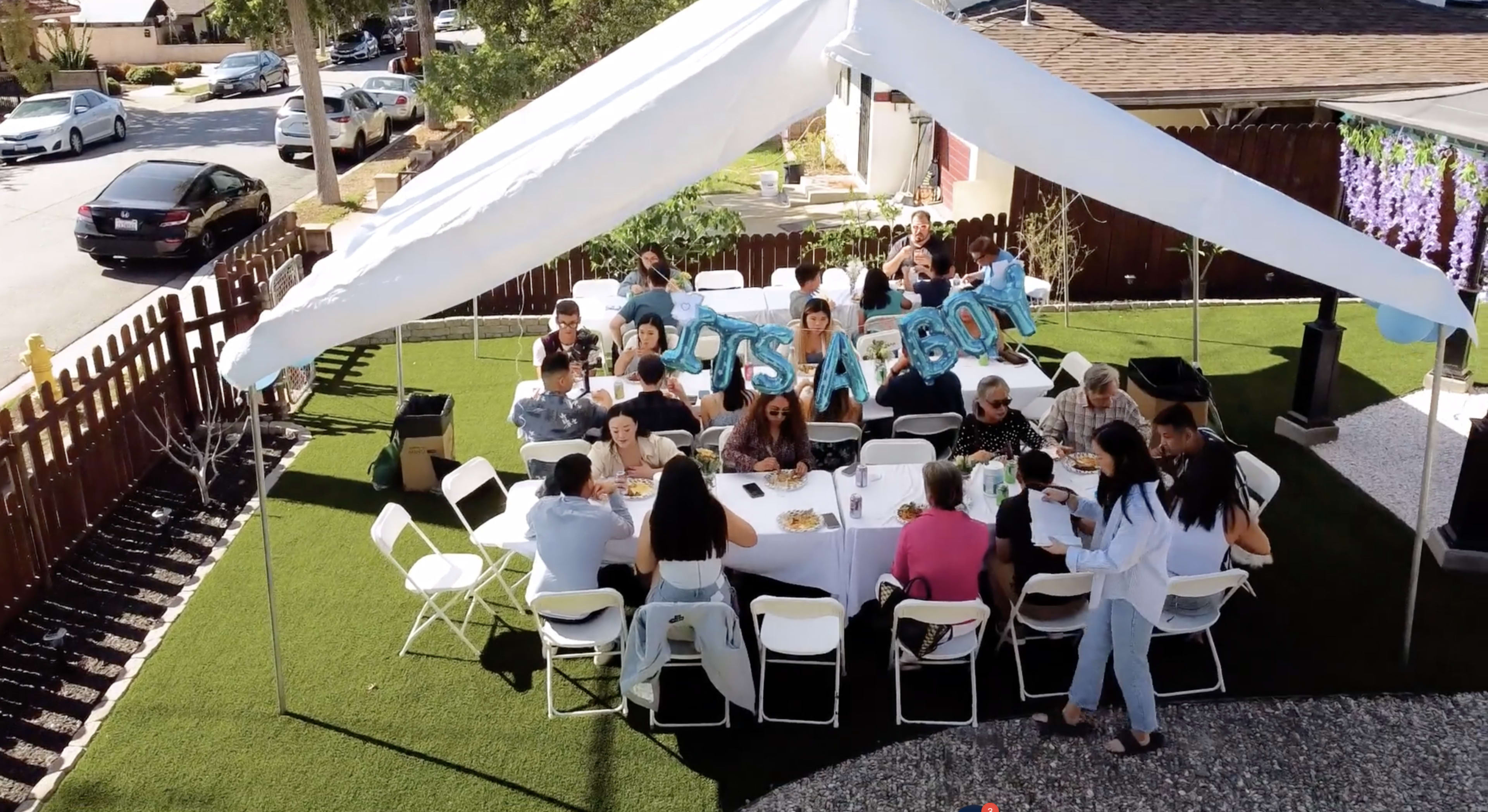 A gathering of people is seated under a white tent with blue "It's a Boy" balloons, celebrating an event in a backyard.