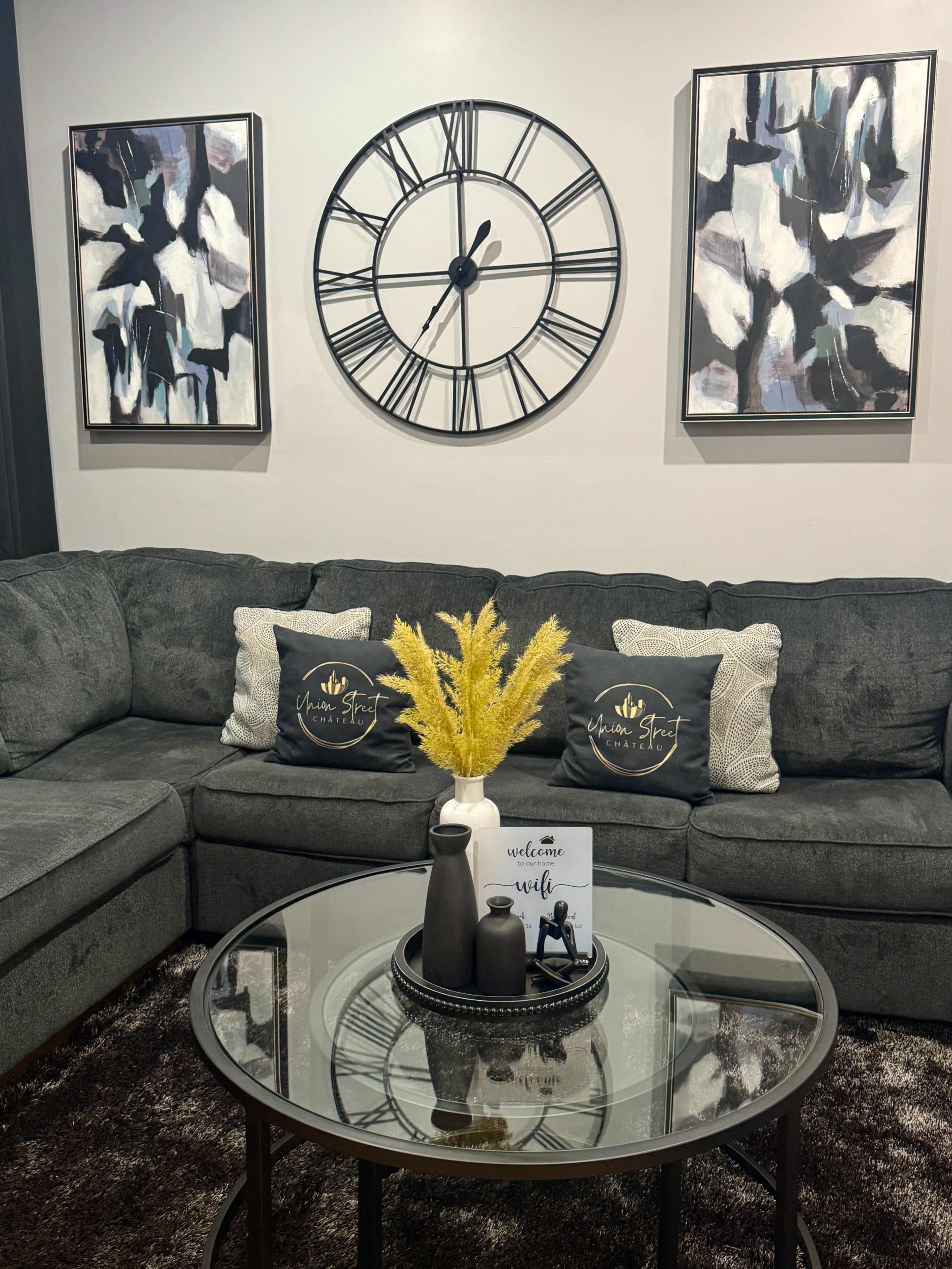 The image shows a modern living room setup featuring a gray sectional sofa, two abstract paintings on the wall, a round glass coffee table, and decorative items including a vase with yellow flowers and a welcome sign.