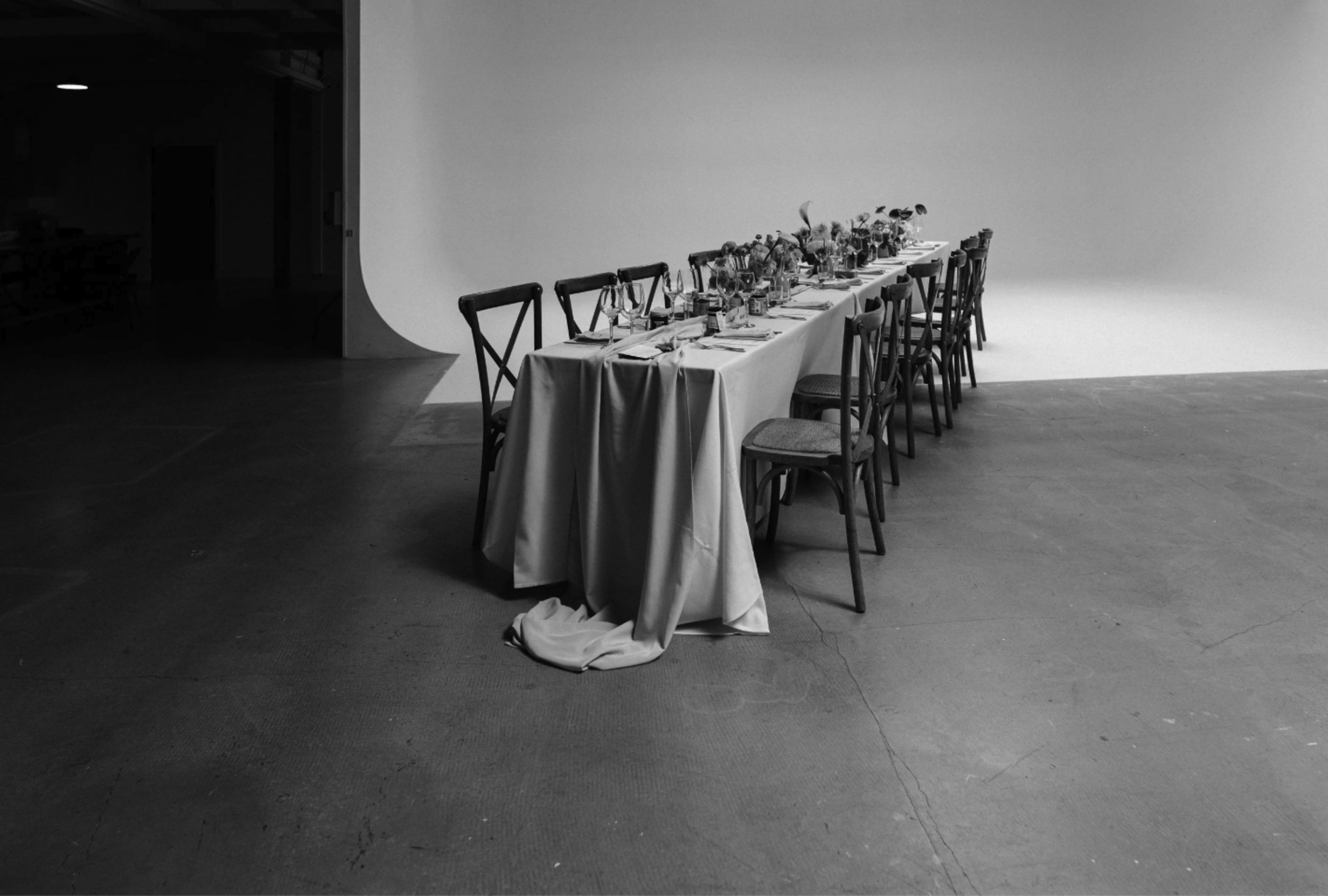 A long dining table with decorations and chairs is set in a minimalist, well-lit room with a plain backdrop.