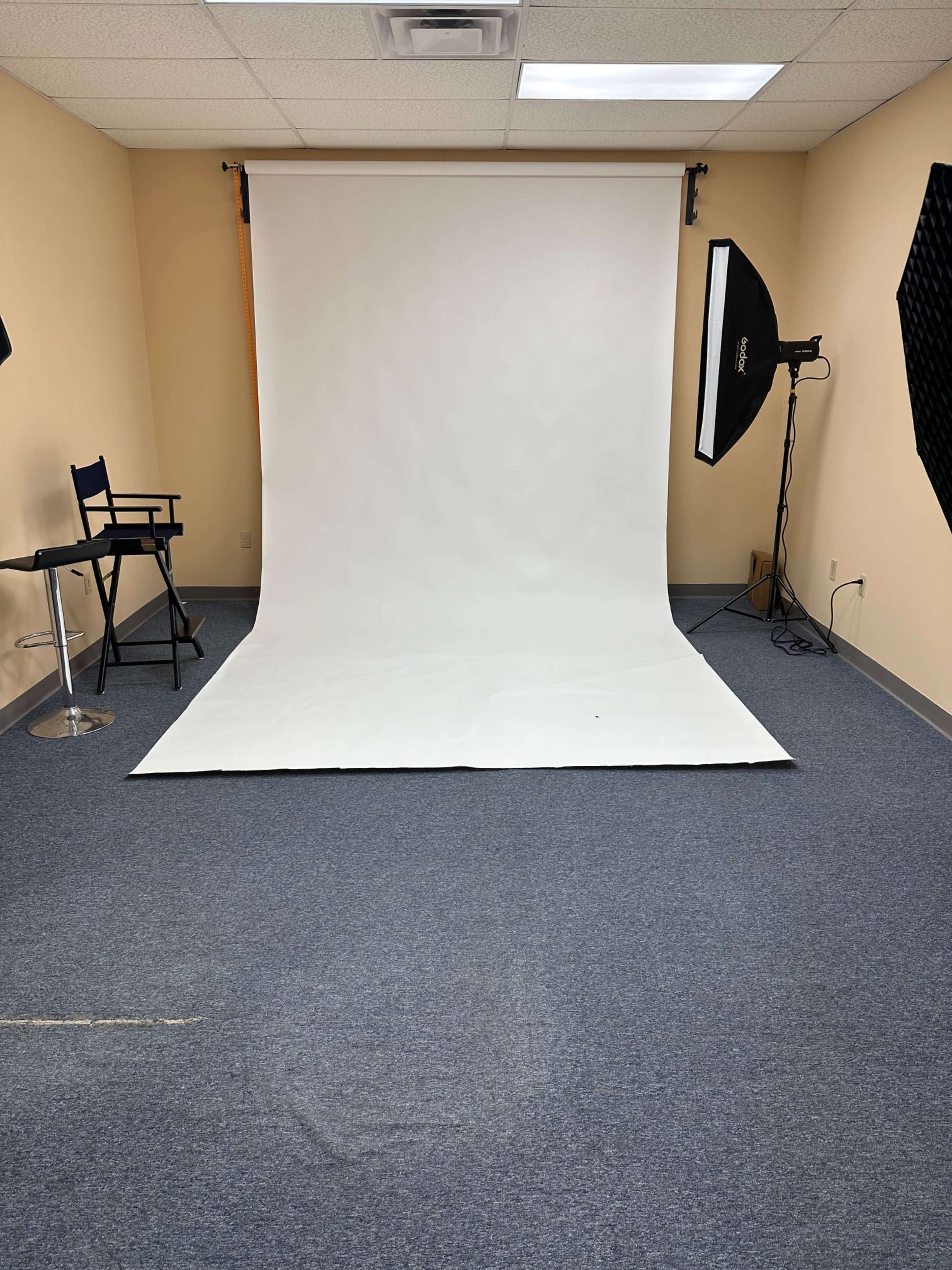 A photography studio with a white backdrop, a chair, and studio lighting equipment set up in a room with gray carpet.