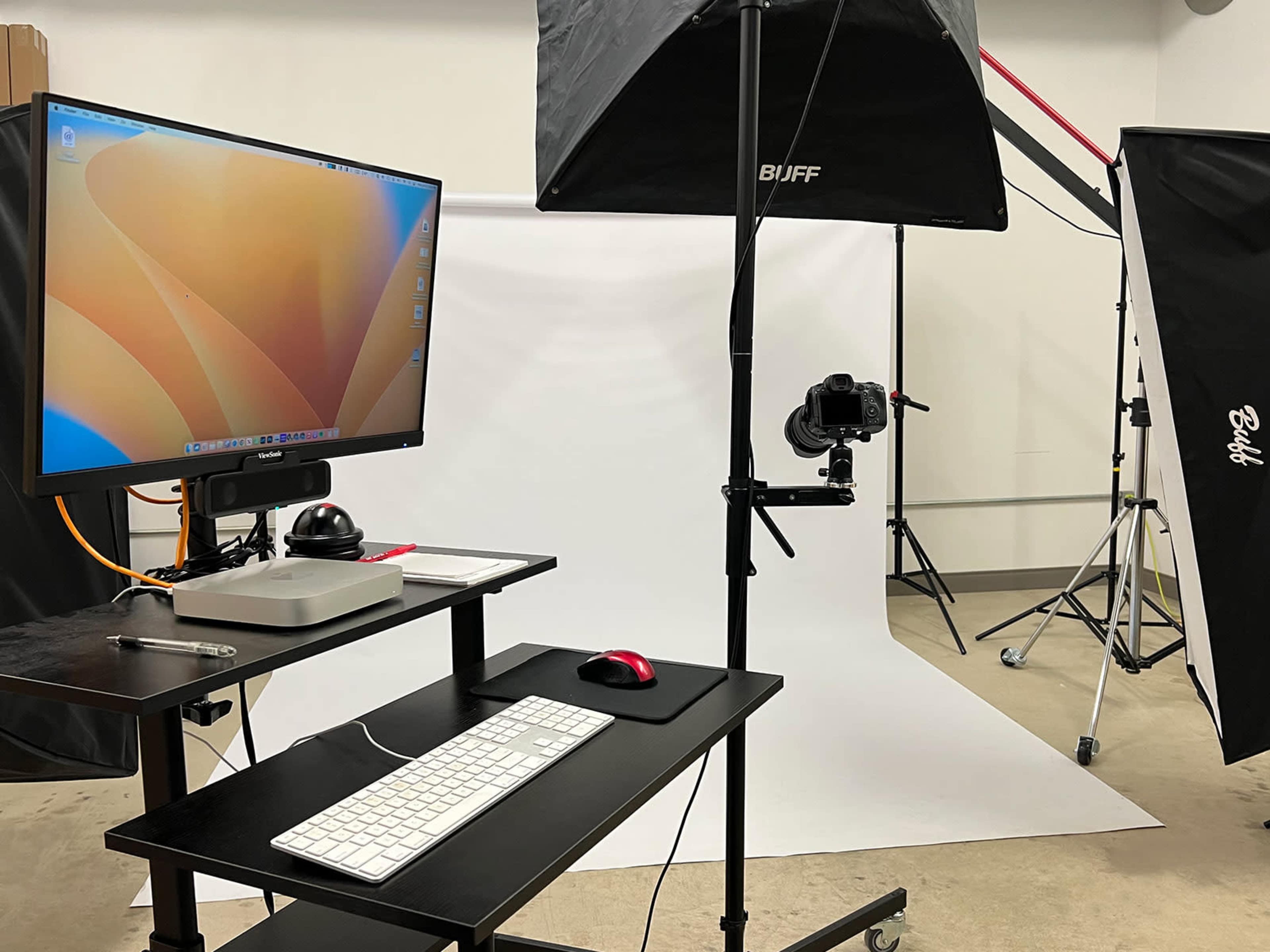 A photography setup featuring a computer monitor, keyboard, and mouse on a desk, along with lighting equipment and a camera facing a white backdrop.