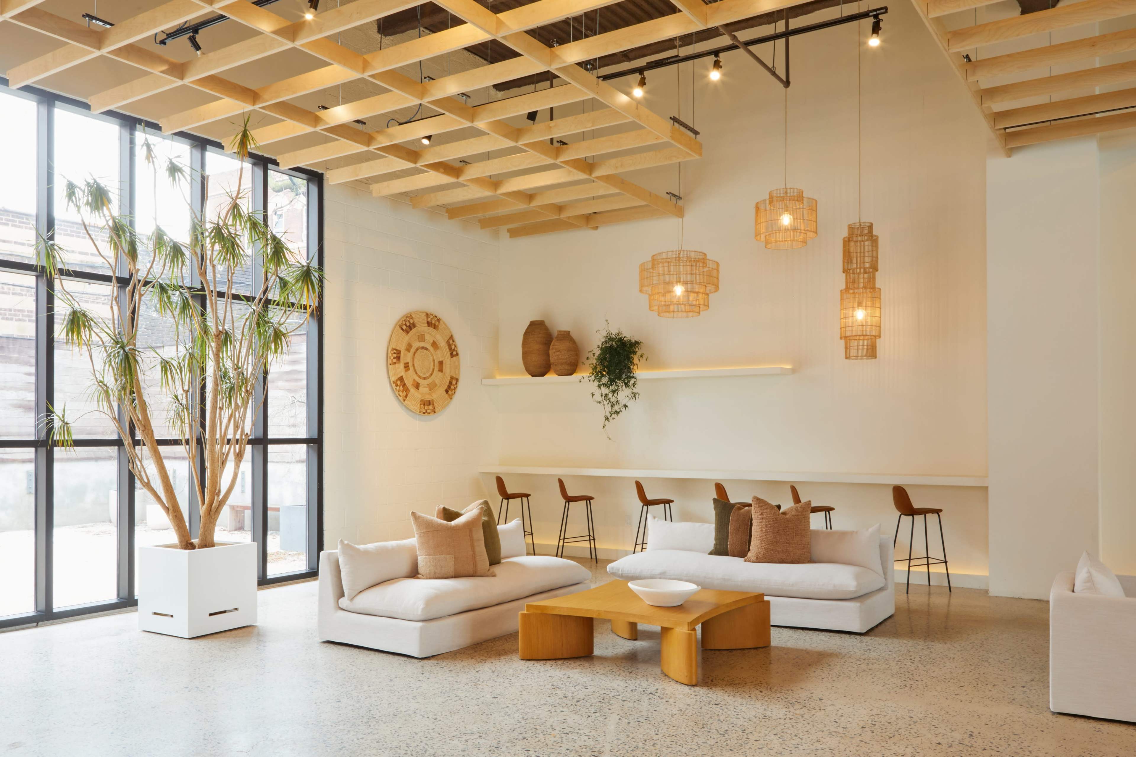 A spacious, modern lounge area features a white sectional sofa, a wooden coffee table, and hanging pendant lights, with large windows allowing natural light to illuminate the space.