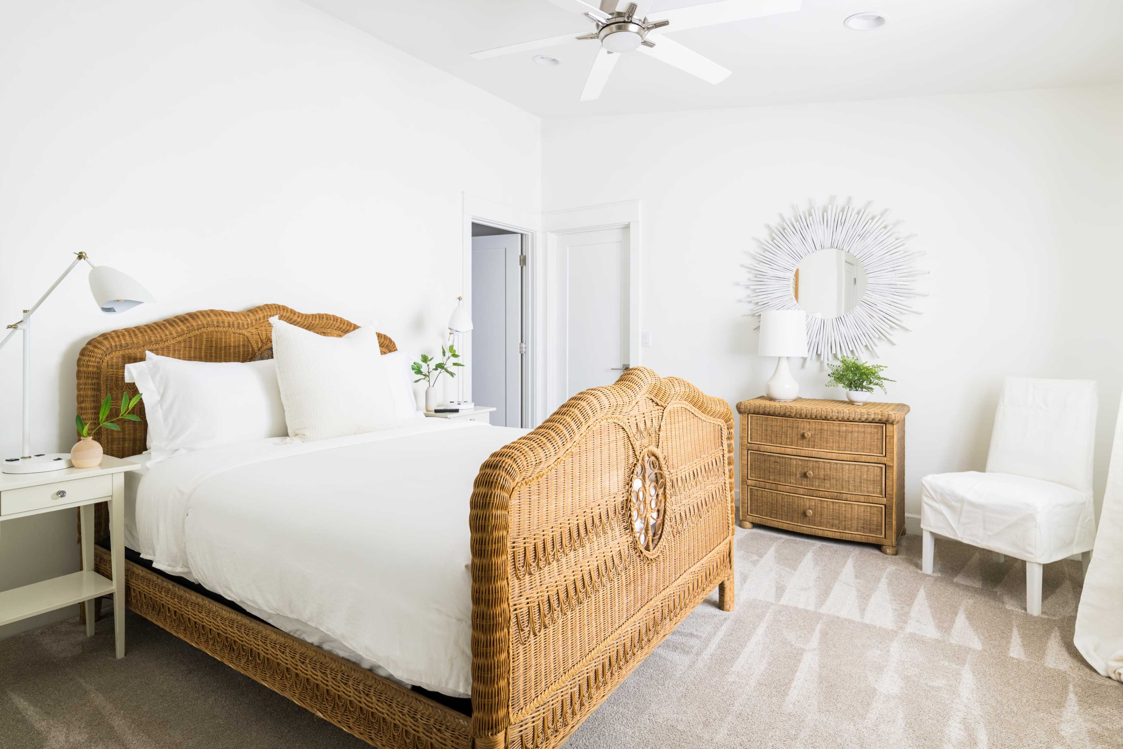 A bright and airy bedroom featuring a wicker bed, a nightstand with a lamp, a mirror, a wooden dresser, and a white chair.