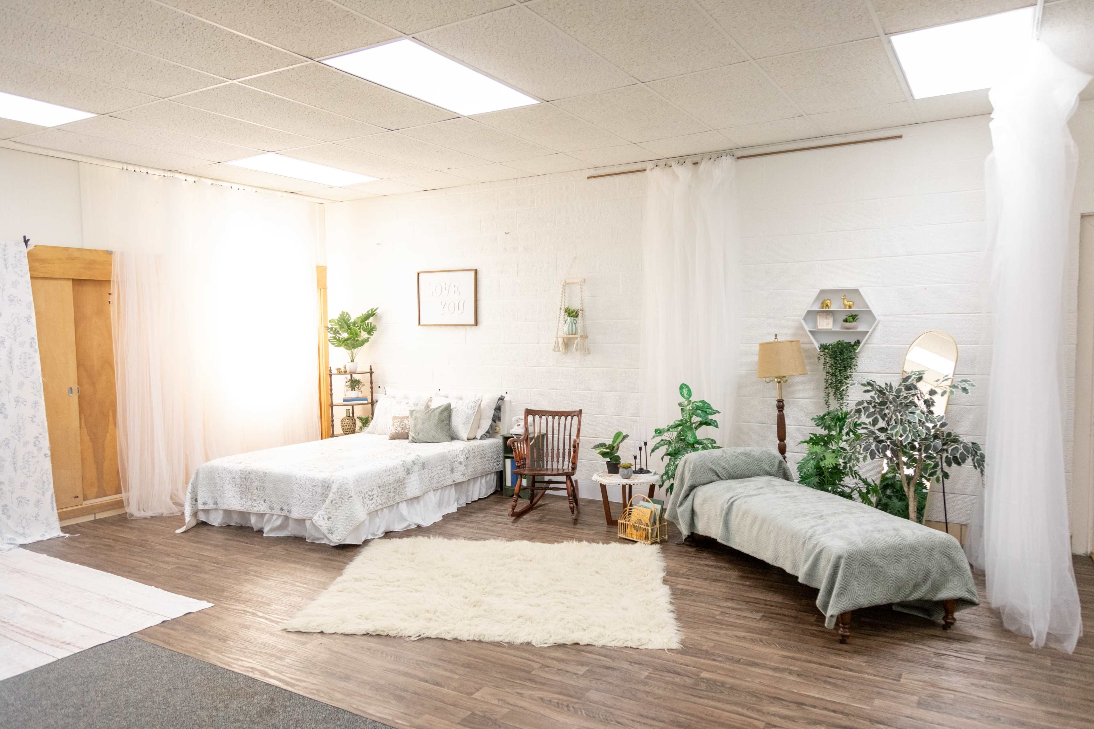 The image shows a styled interior of a room featuring two beds, one with a decorative bedspread and the other with a simple blanket, surrounded by various plants and a lamp.