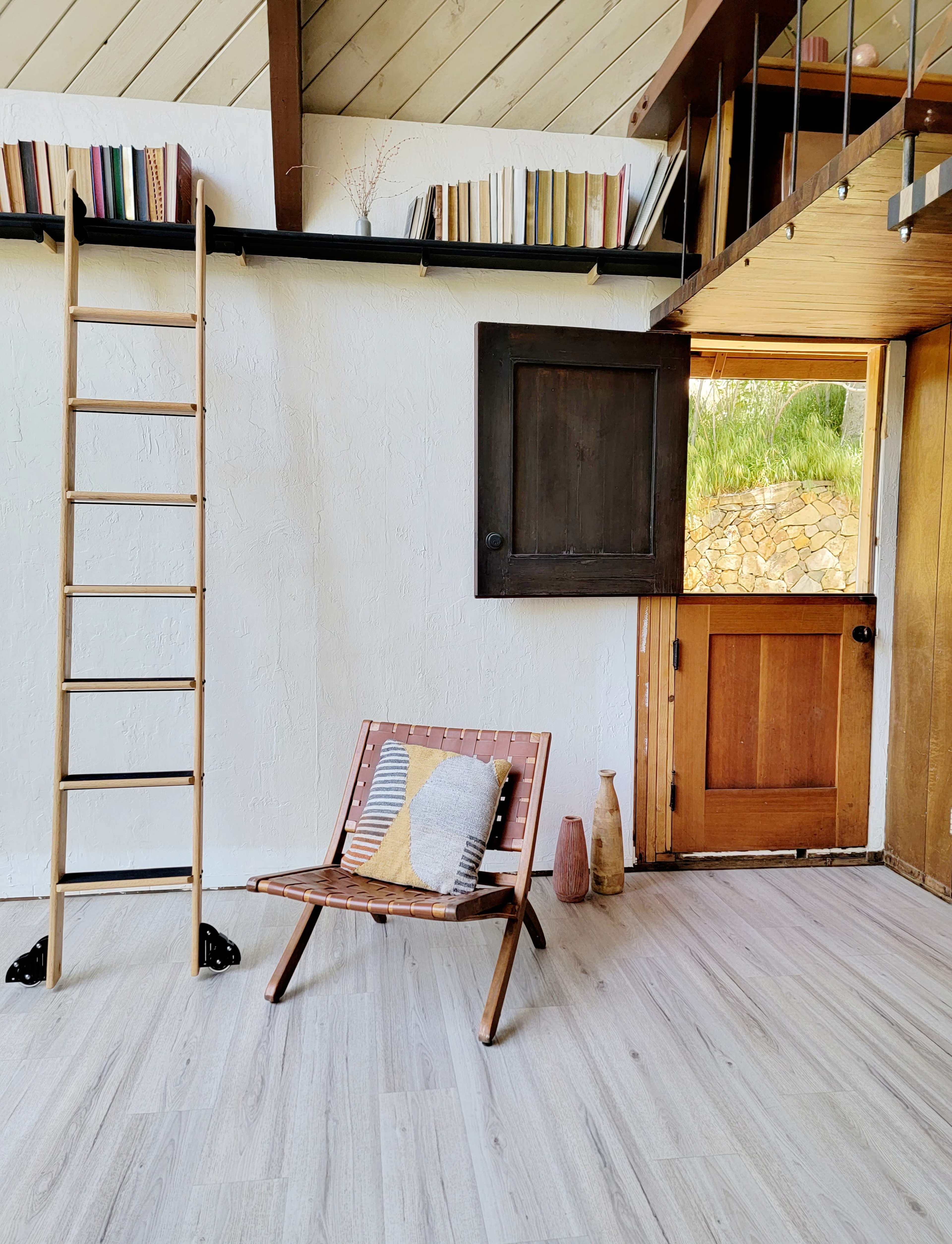 The image shows a minimalist interior featuring a wooden ladder, a slatted chair with a cushion, and an open door leading to an outdoor area.