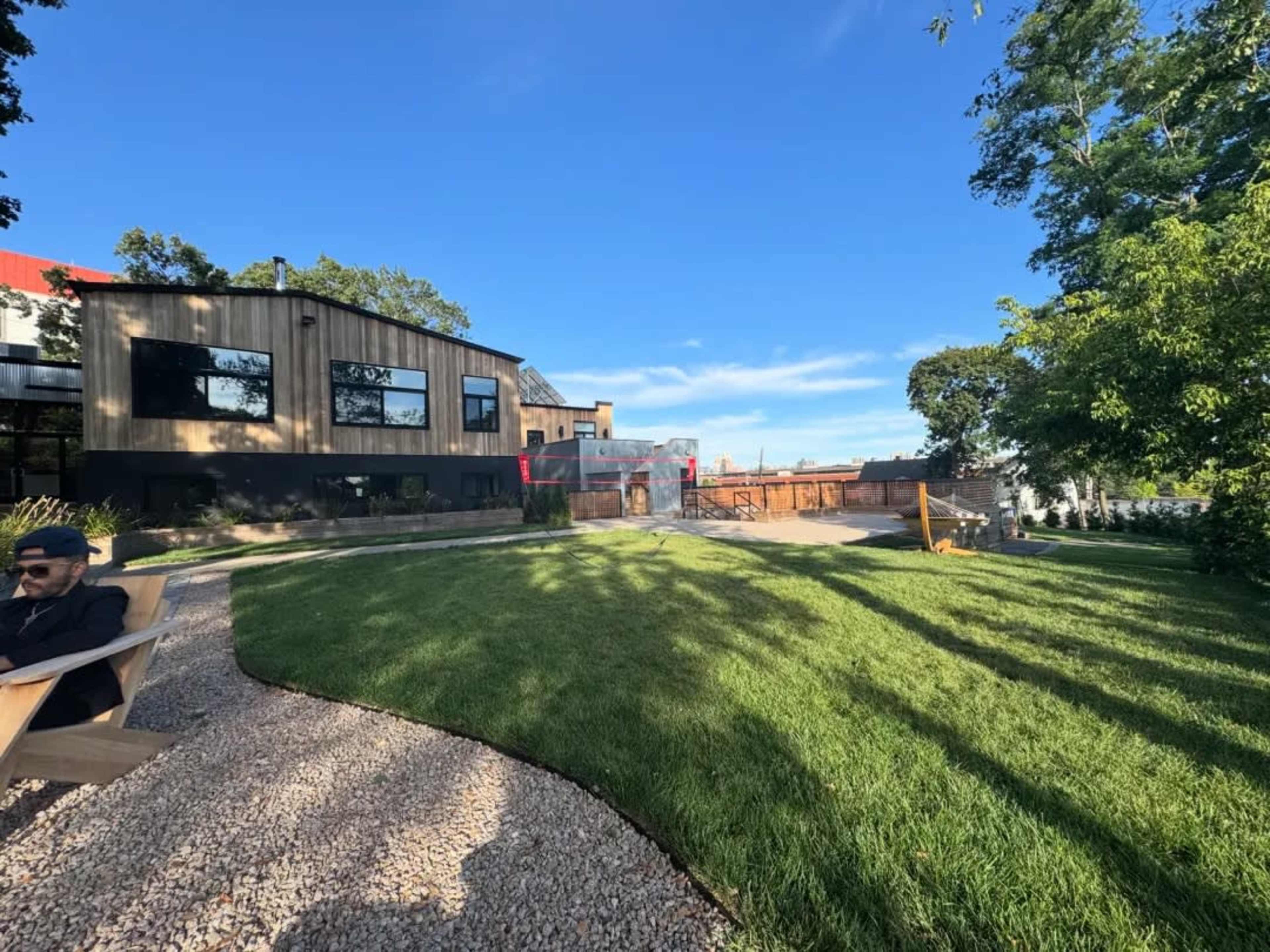 A well-maintained grassy area surrounds a modern building with a mix of wood and glass exterior, alongside gravel pathways and trees.