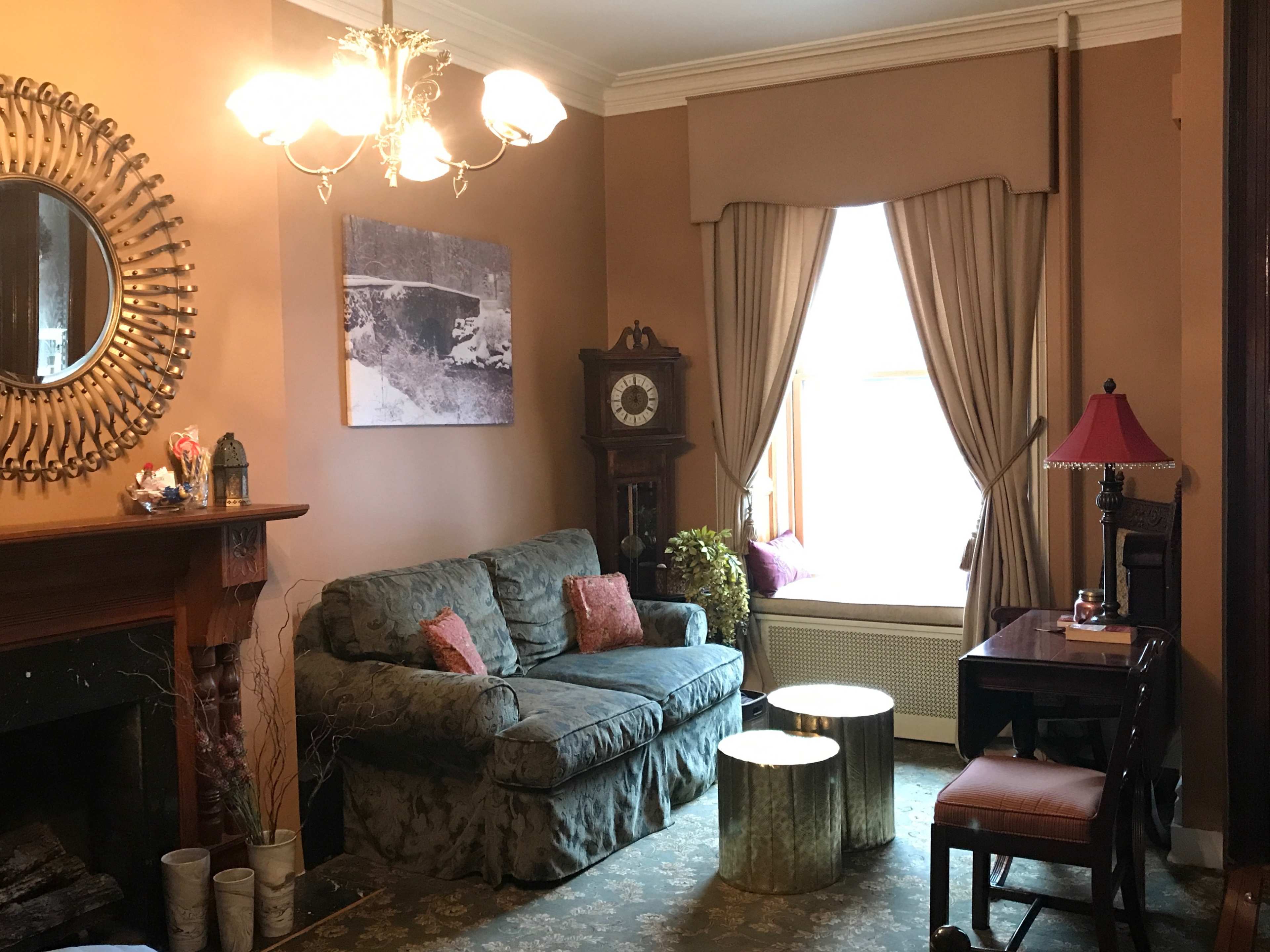 A living room with a green sofa, a round mirror over the fireplace, an antique clock, a desk, and a window with beige curtains.