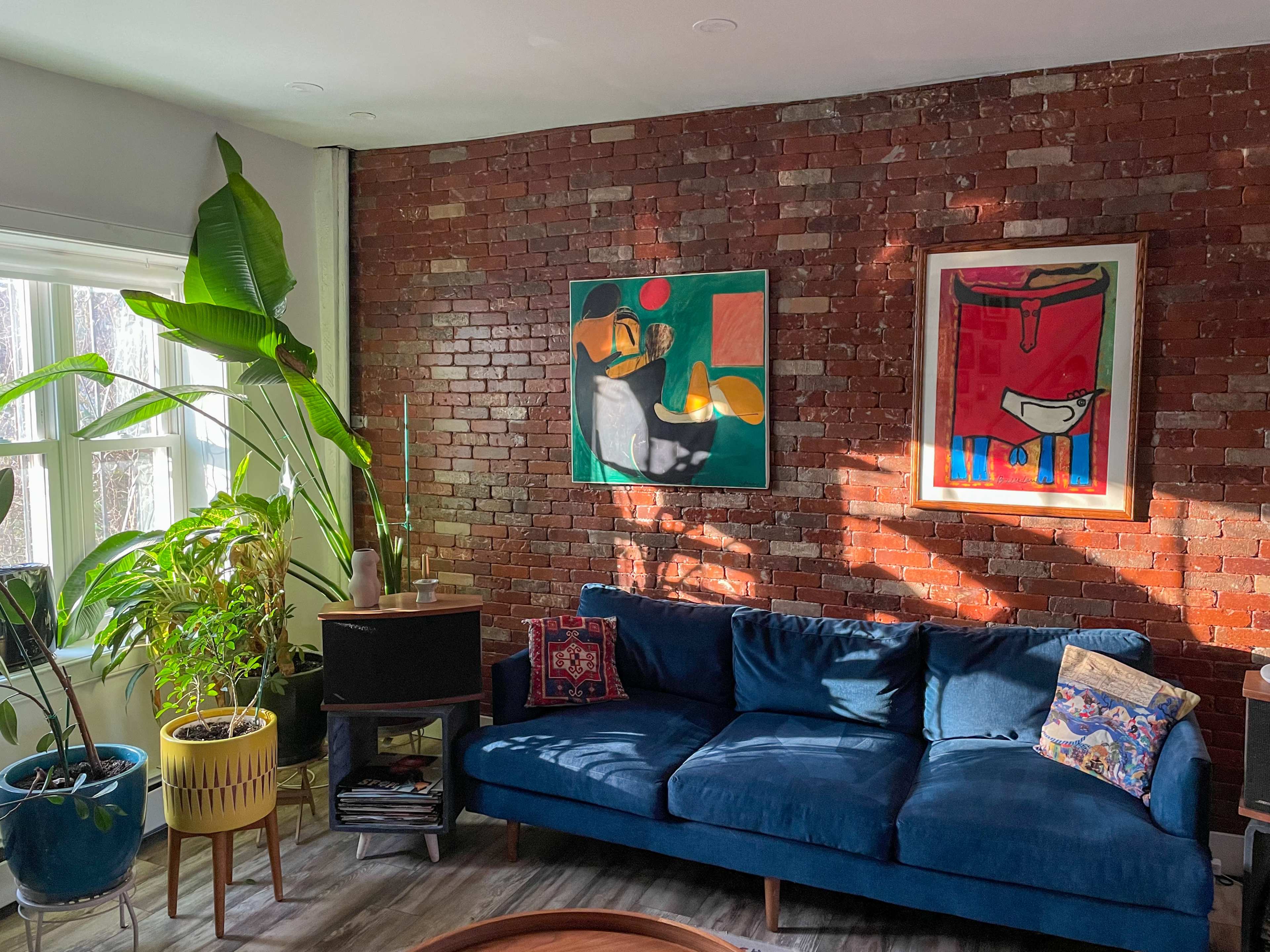 A blue velvet sofa is positioned against a brick wall, adorned with two colorful abstract paintings and surrounded by various indoor plants.