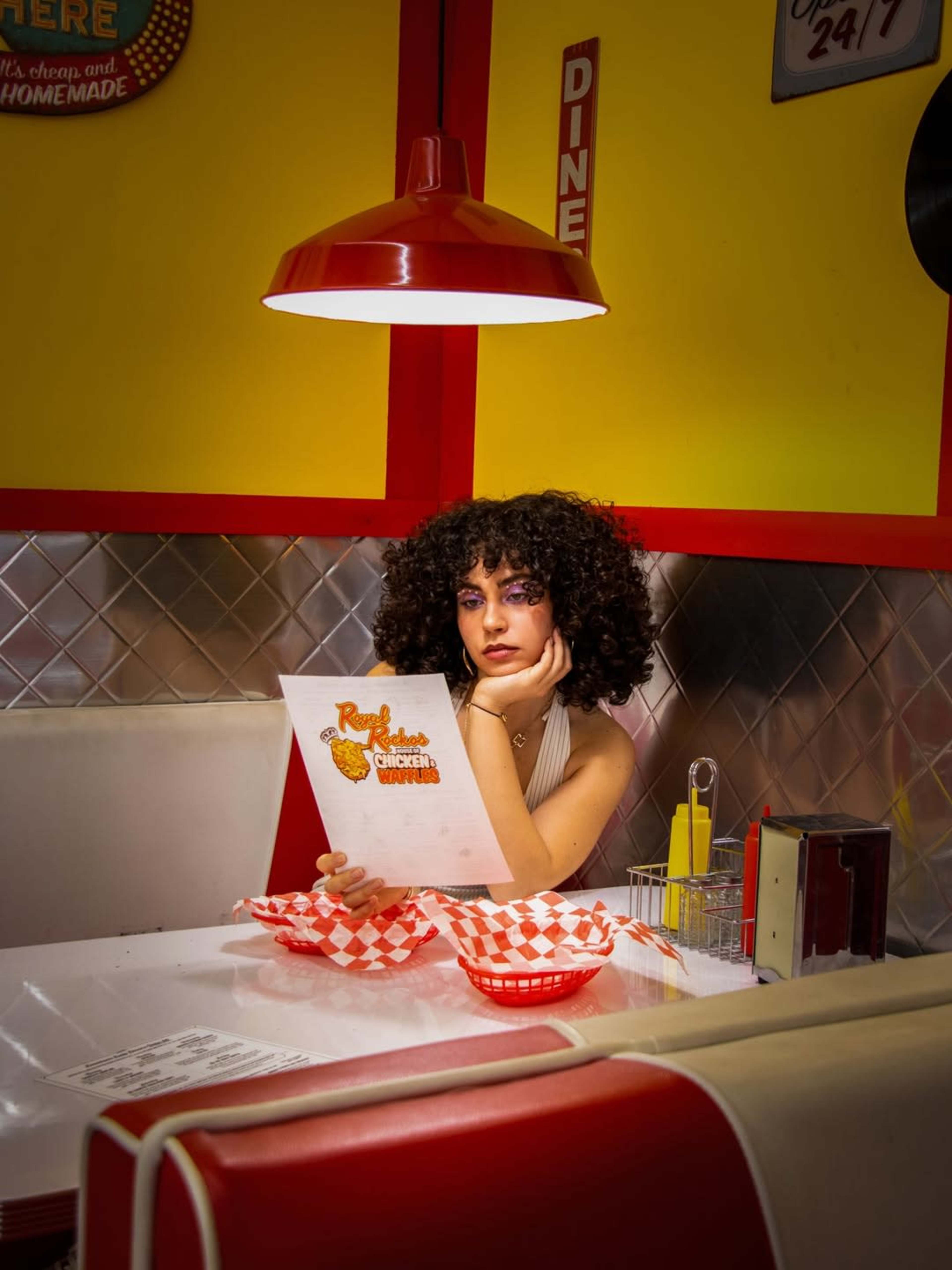 A woman with curly hair sits at a retro diner table, examining a menu beneath a red pendant light.