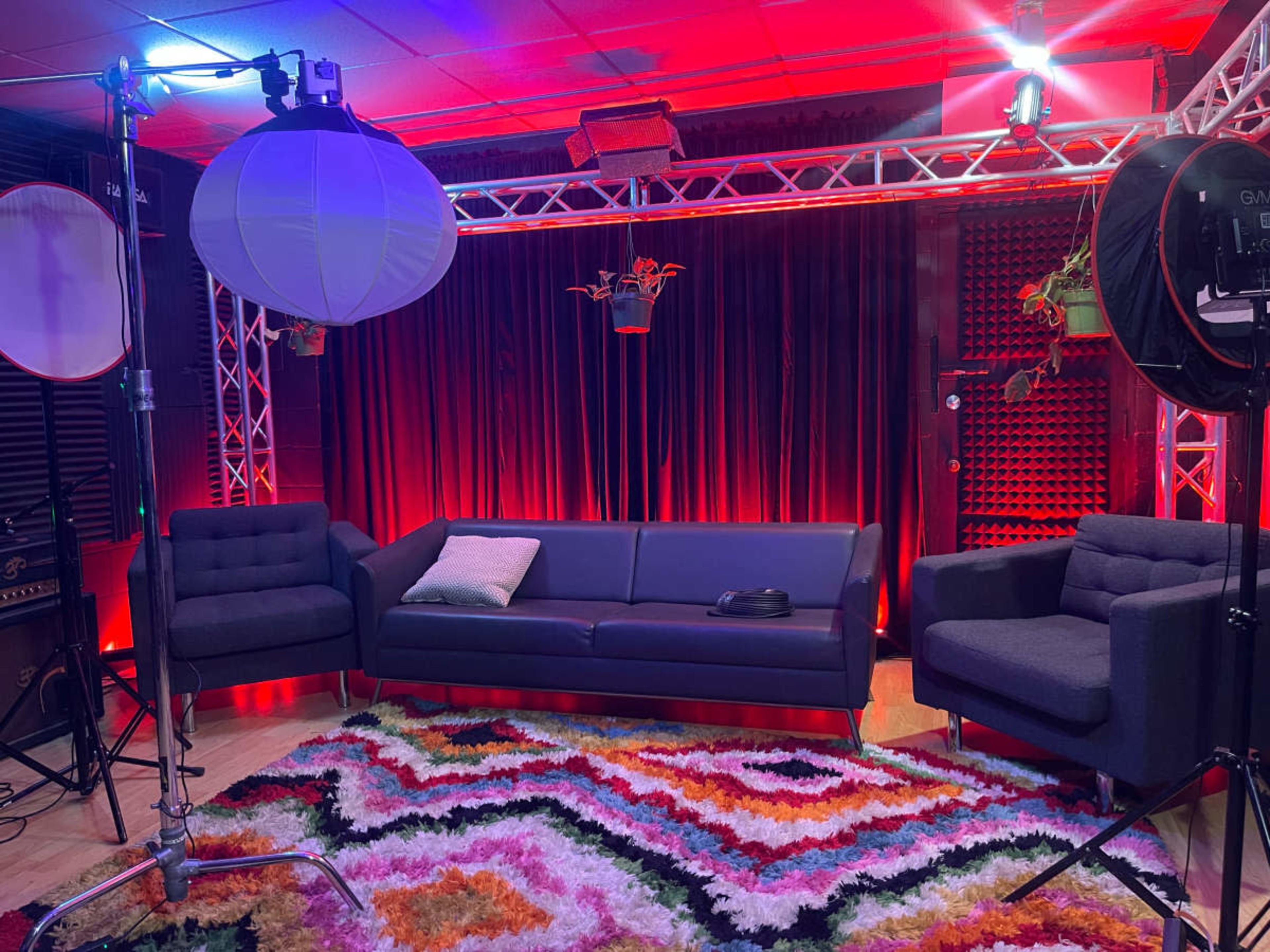 The image shows a brightly lit studio setup with a dark sofa, colorful rug, and various lighting equipment arranged around a backdrop of red curtains.