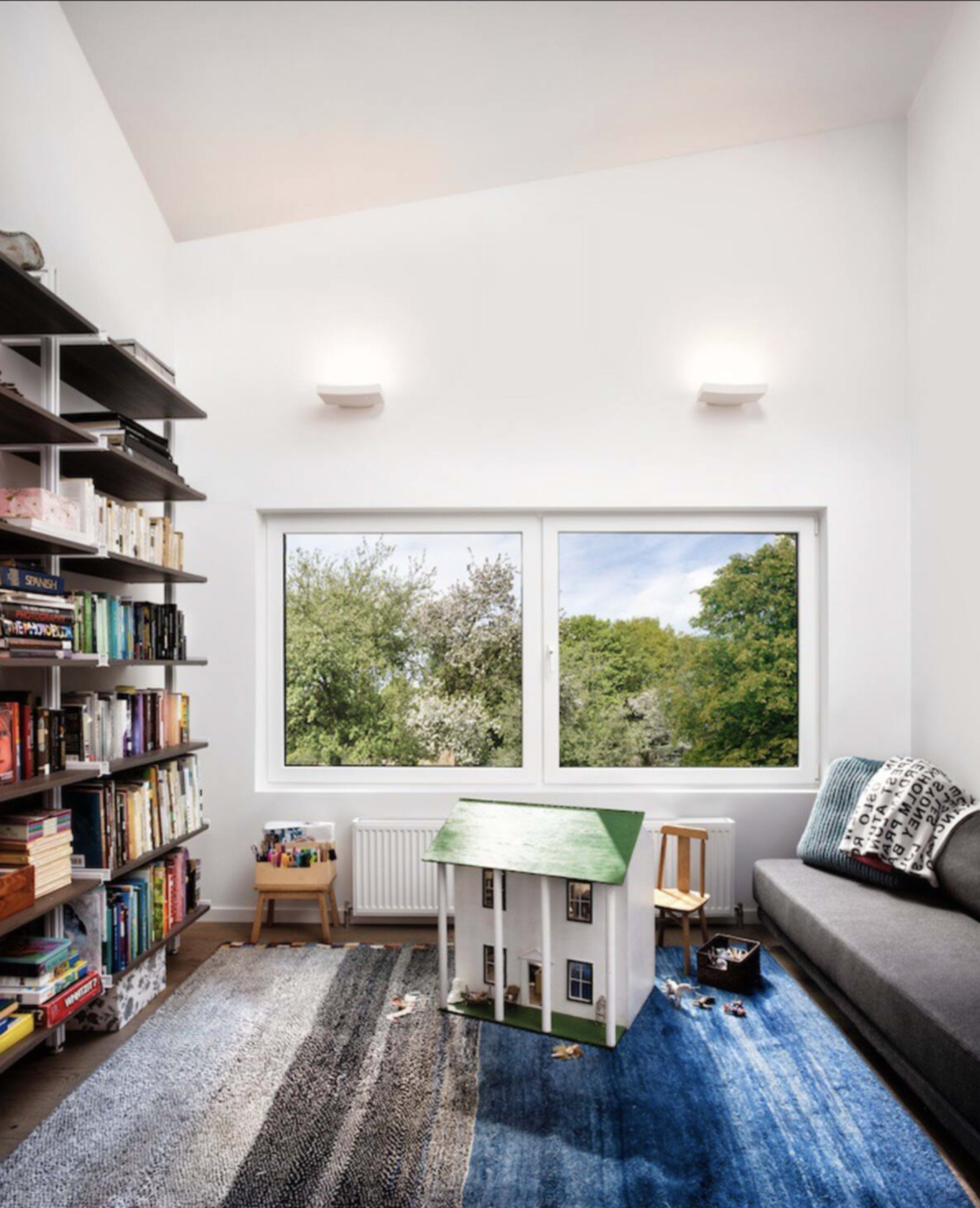 A well-lit room features a bookshelf filled with books, a grey sofa, and a toy house on a patterned rug, with large windows overlooking greenery.