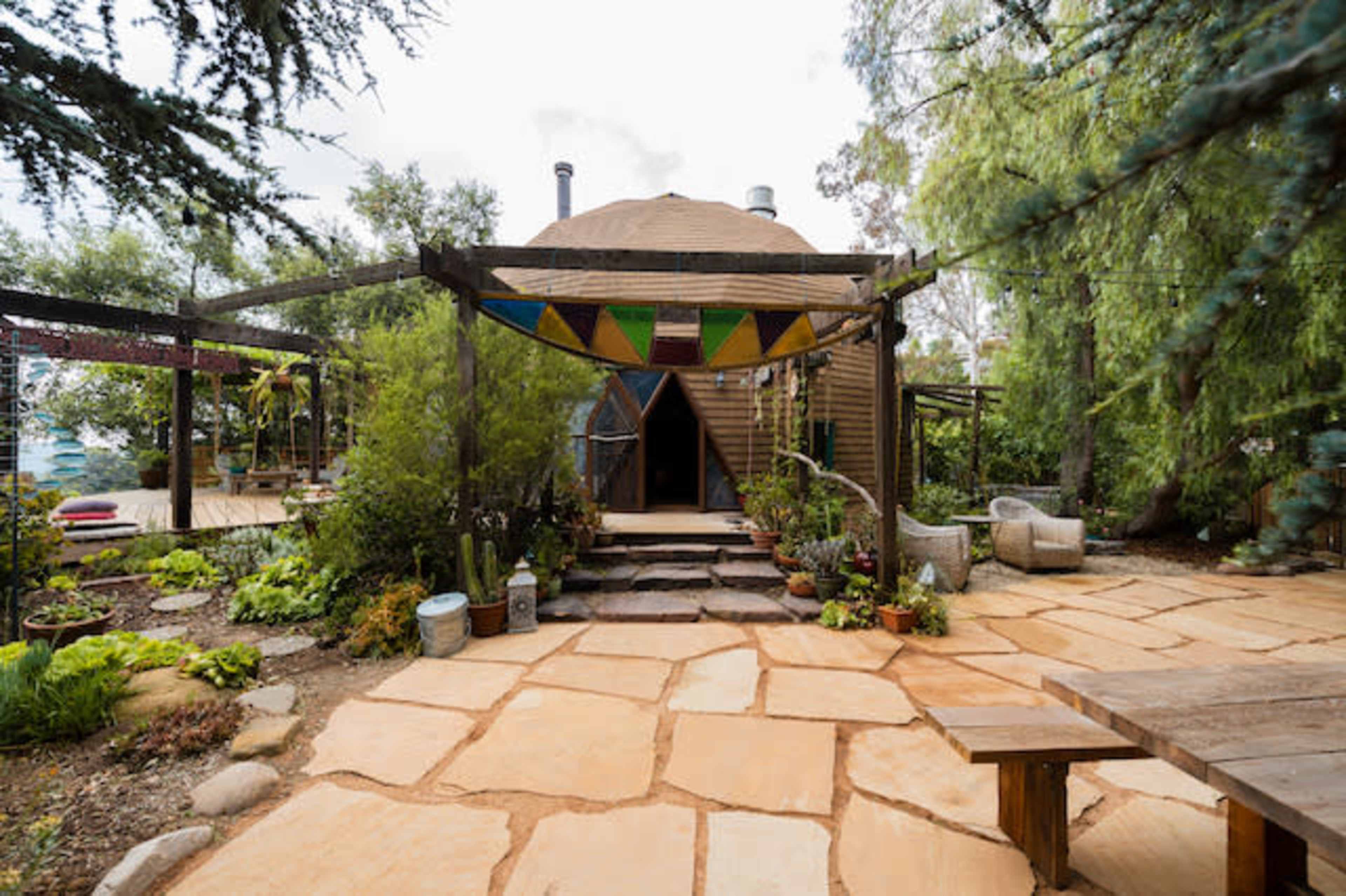 A round, wooden structure with a distinct entrance is surrounded by greenery and a stone pathway, leading to an outdoor seating area.