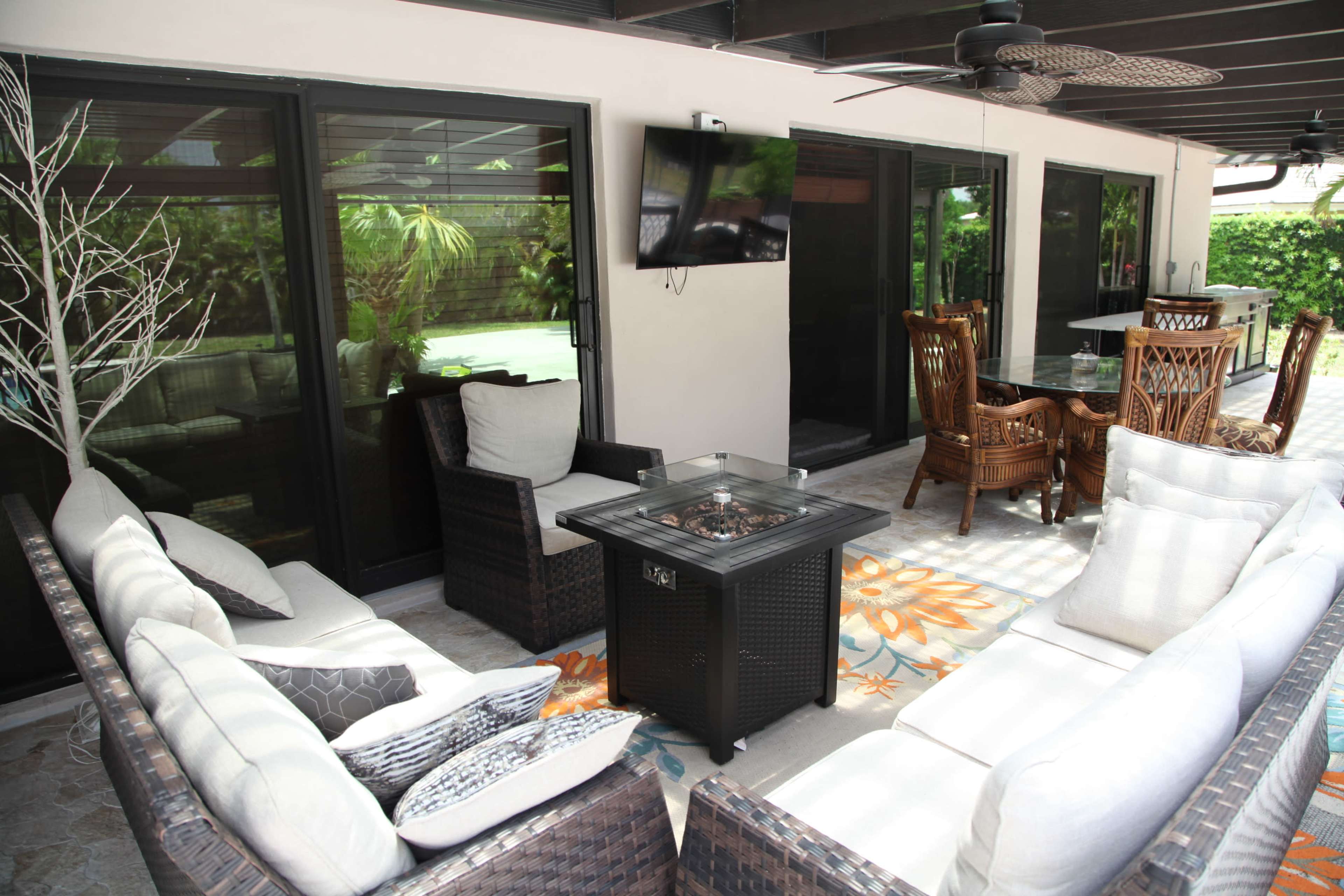 A covered patio area with wicker furniture arranged around a fire pit, a television mounted on the wall, and a dining area in the background.