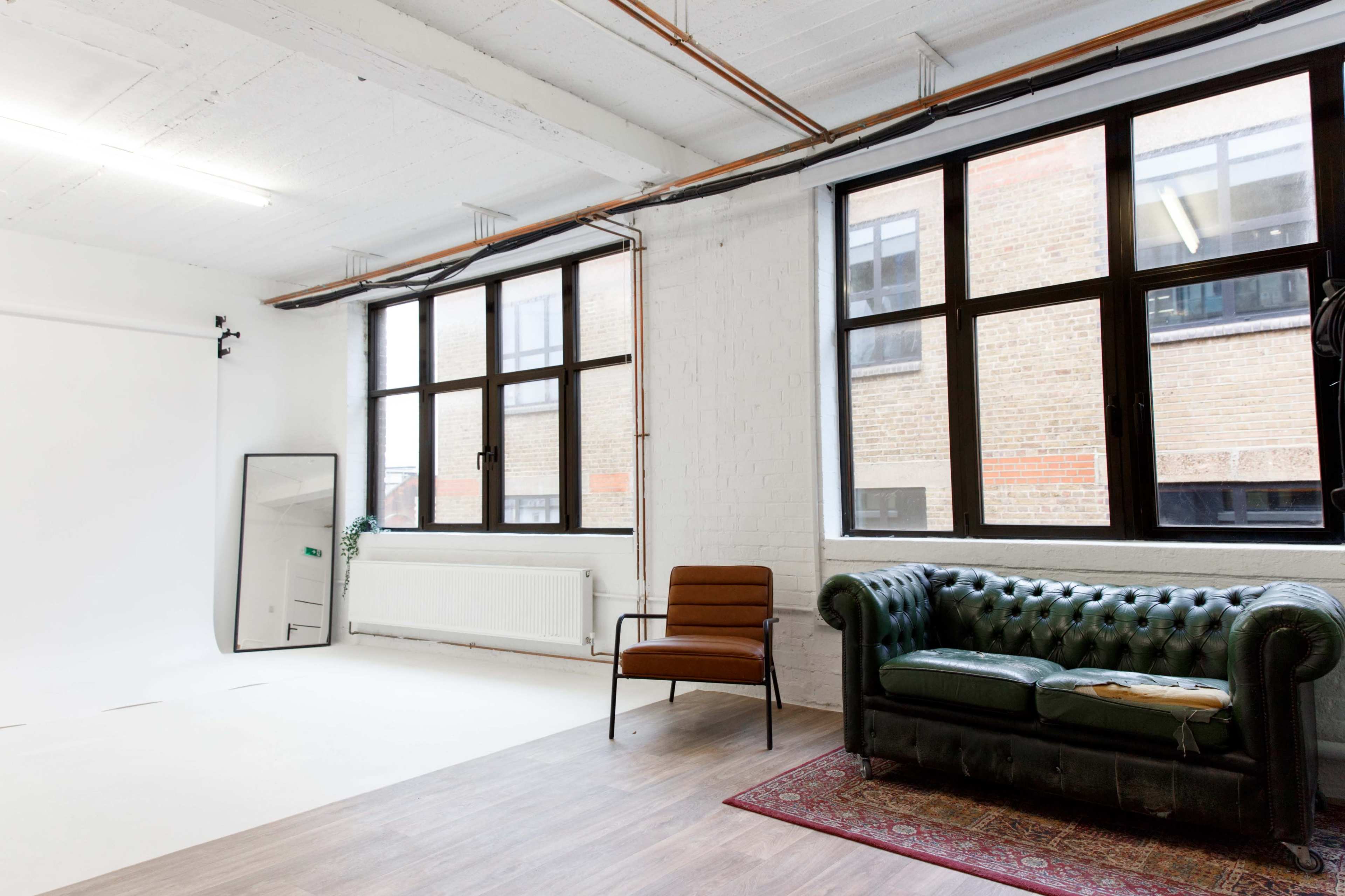 A spacious room with large windows, a green leather sofa, a brown chair, and a mirror against a white wall.