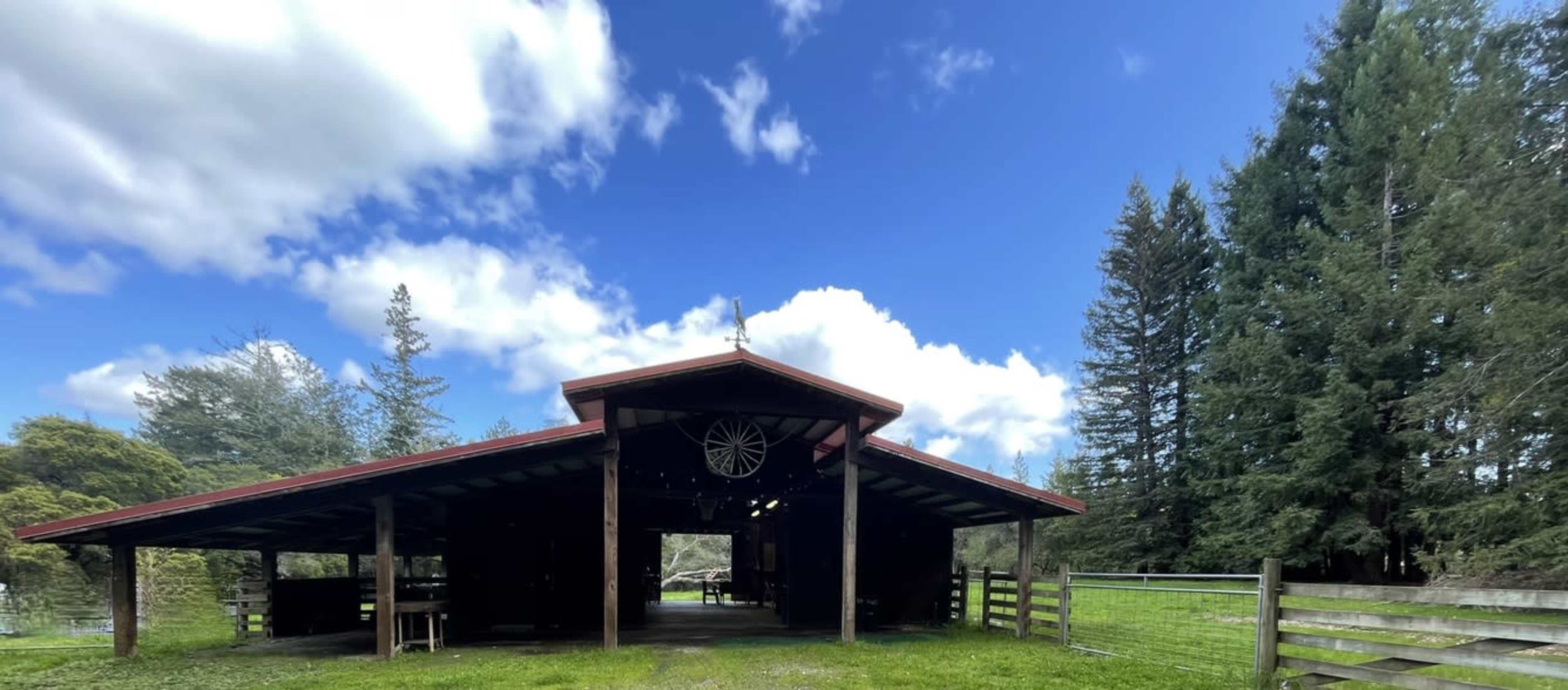 A large, open barn with a red roof and wooden beams stands under a blue sky adorned with fluffy clouds, surrounded by green grass and trees.