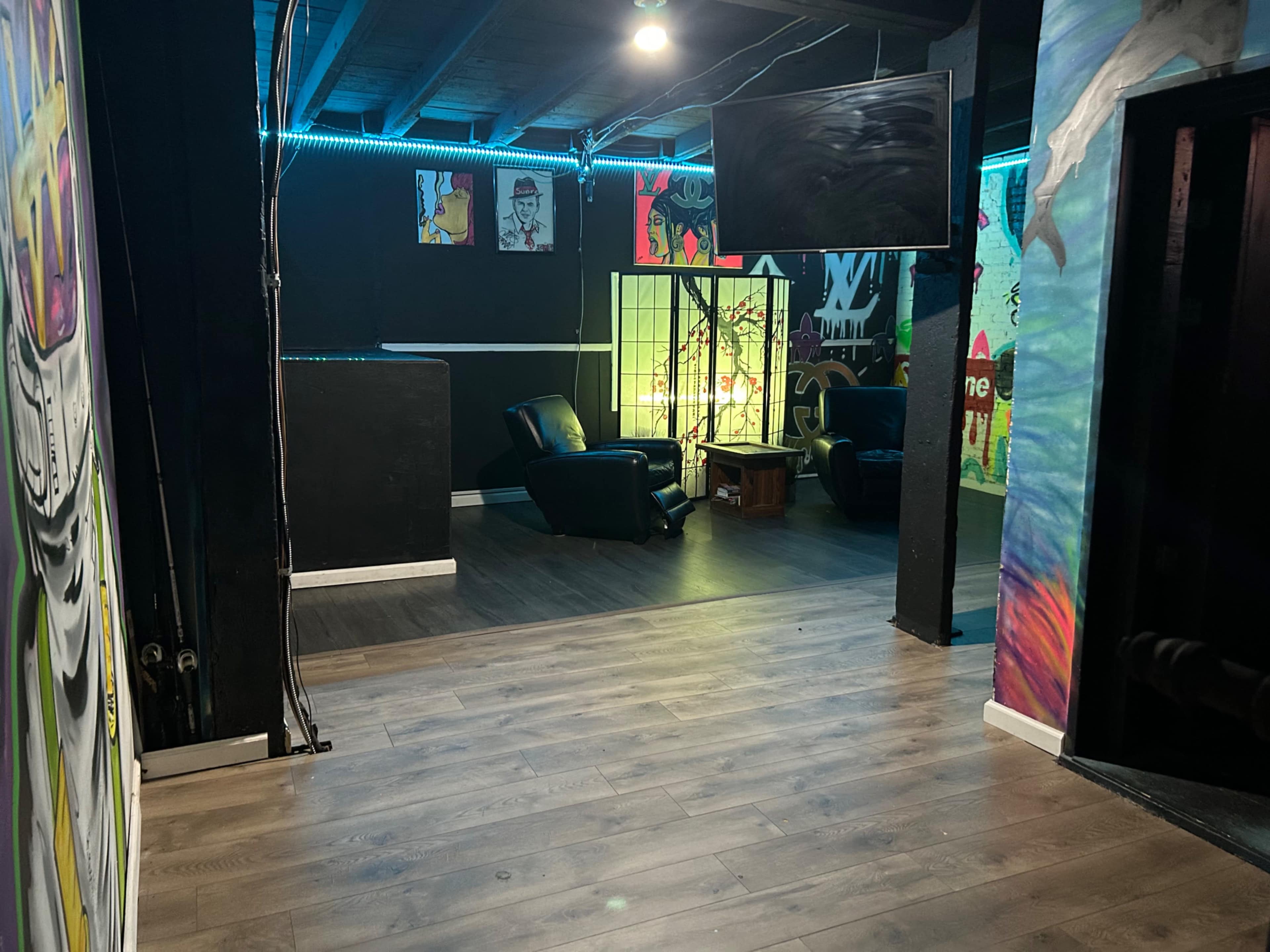 A dimly lit room featuring a television on a stand, two leather chairs, and colorful murals on the walls.