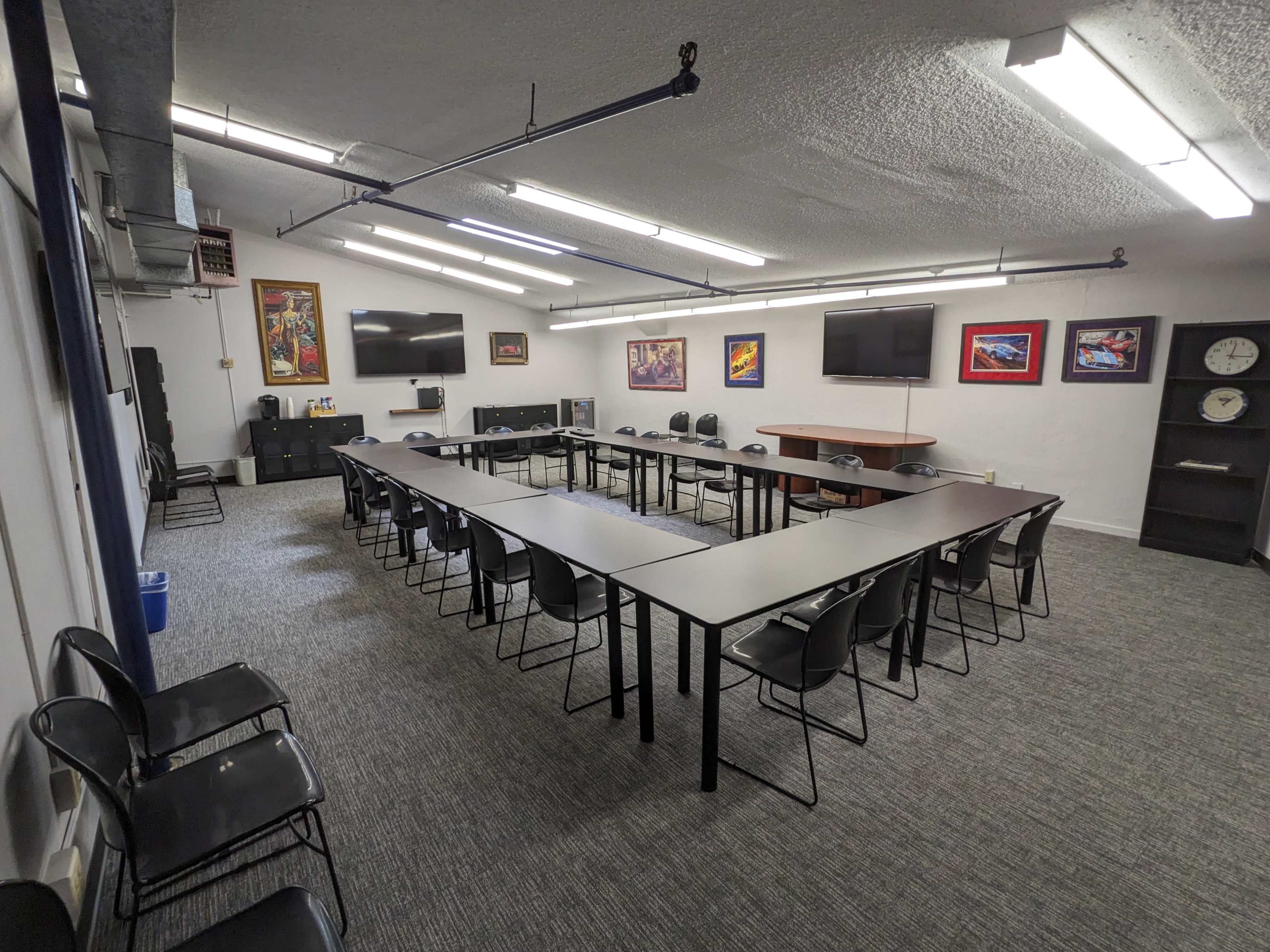 The image shows a spacious meeting room with several long tables arranged in a U-shape, surrounded by black chairs, and equipped with multiple screens on the walls.