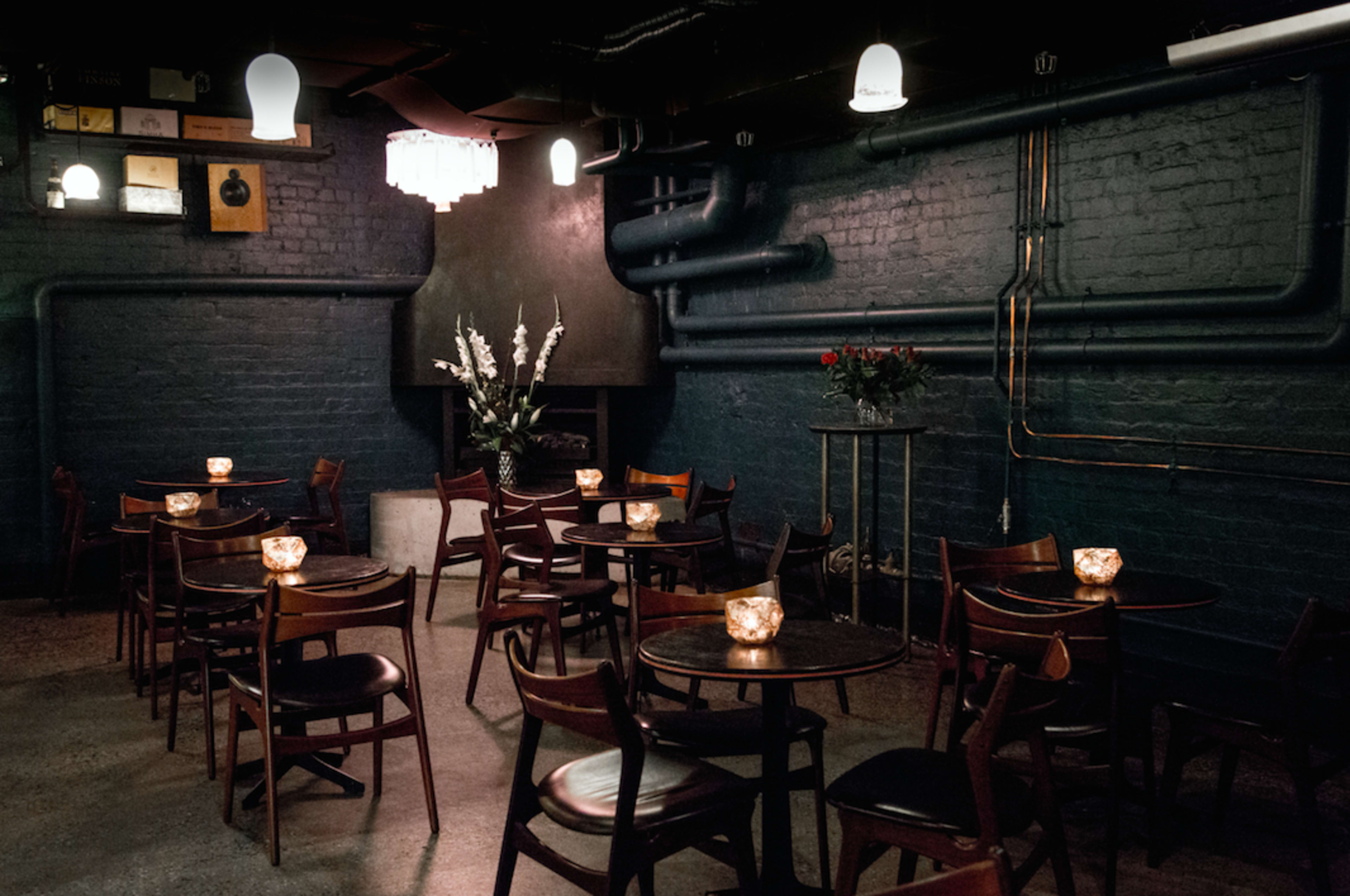 The image shows a dimly lit restaurant interior with wooden tables, chairs, and decorative candles on each table against a backdrop of exposed pipes and floral arrangements.