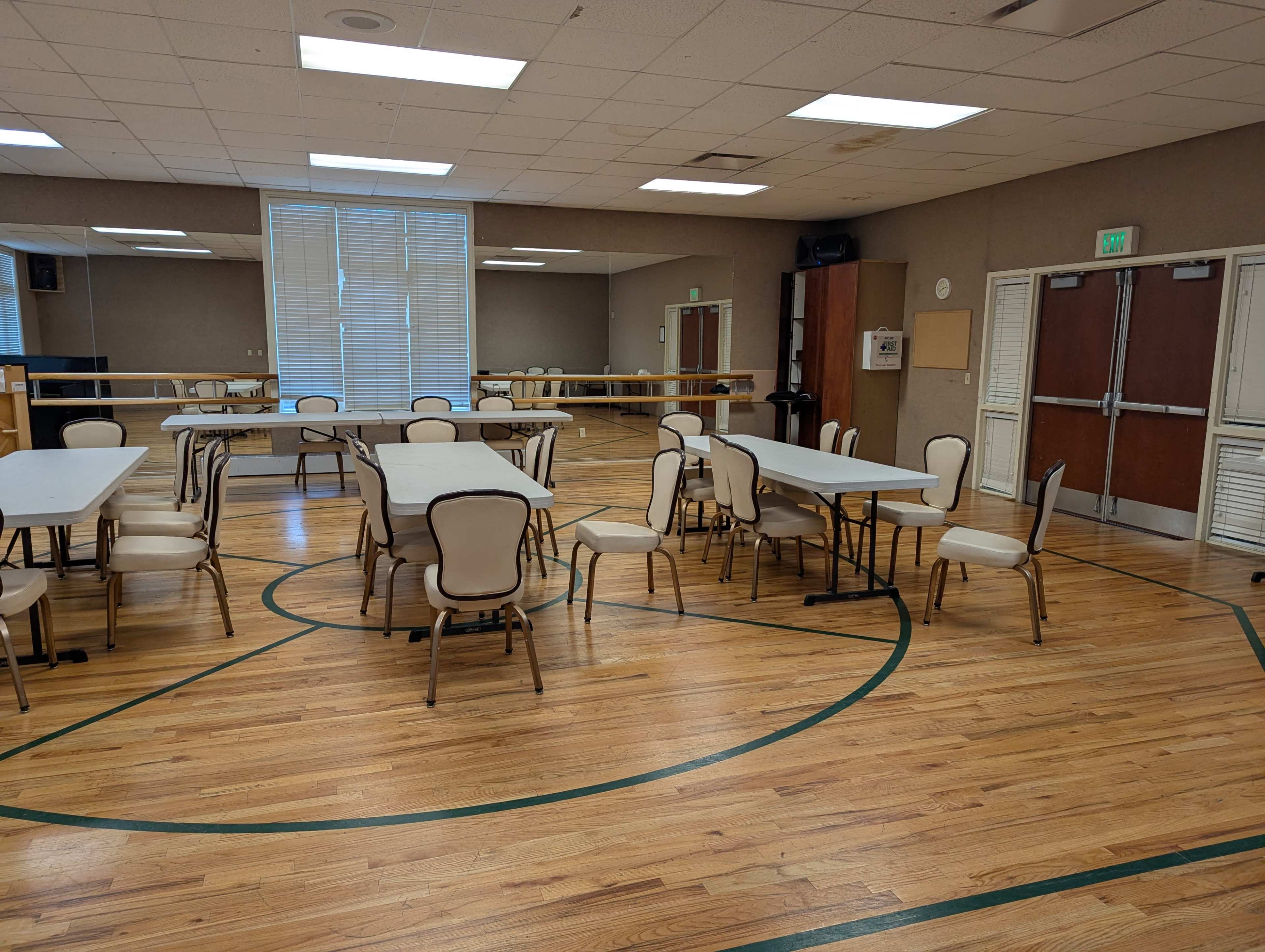 The image shows a spacious room with wooden floors, several round and rectangular tables, and chairs arranged around them, along with mirrors and a ballet bar along one wall.