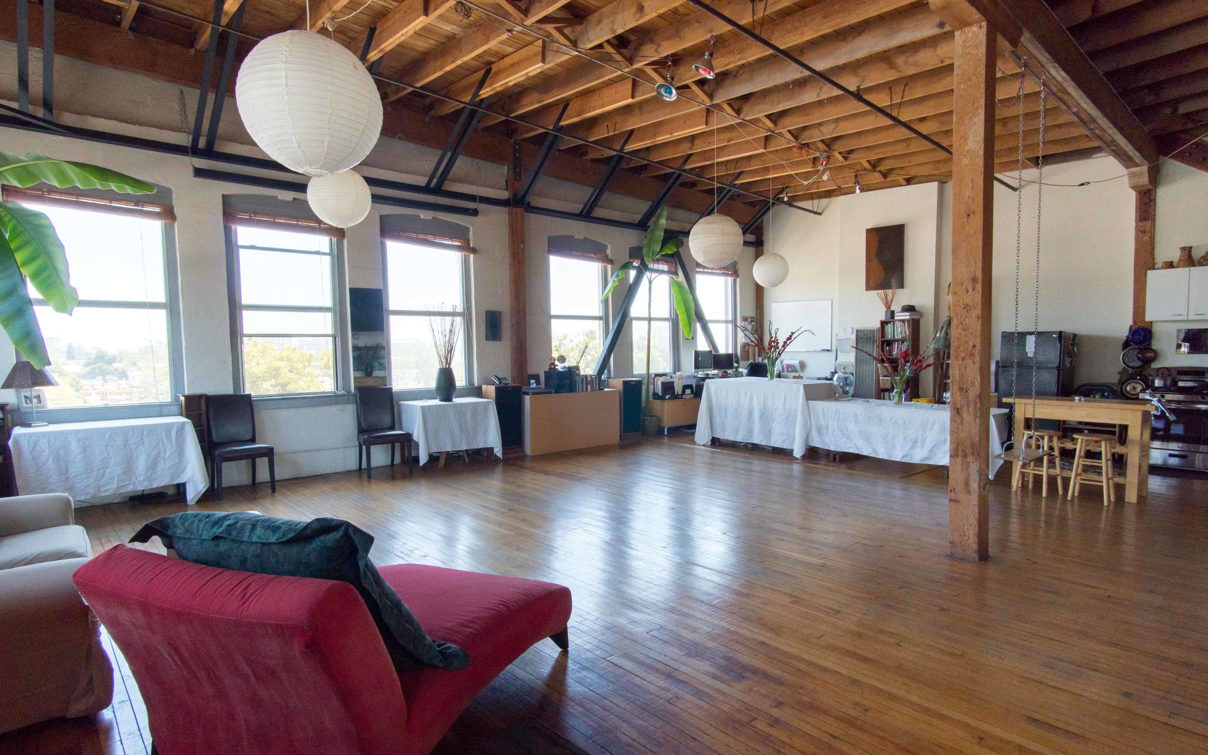 A spacious room with wooden floors, high ceilings, and large windows, featuring minimal furniture including a red armchair, white tables, and various decorations.