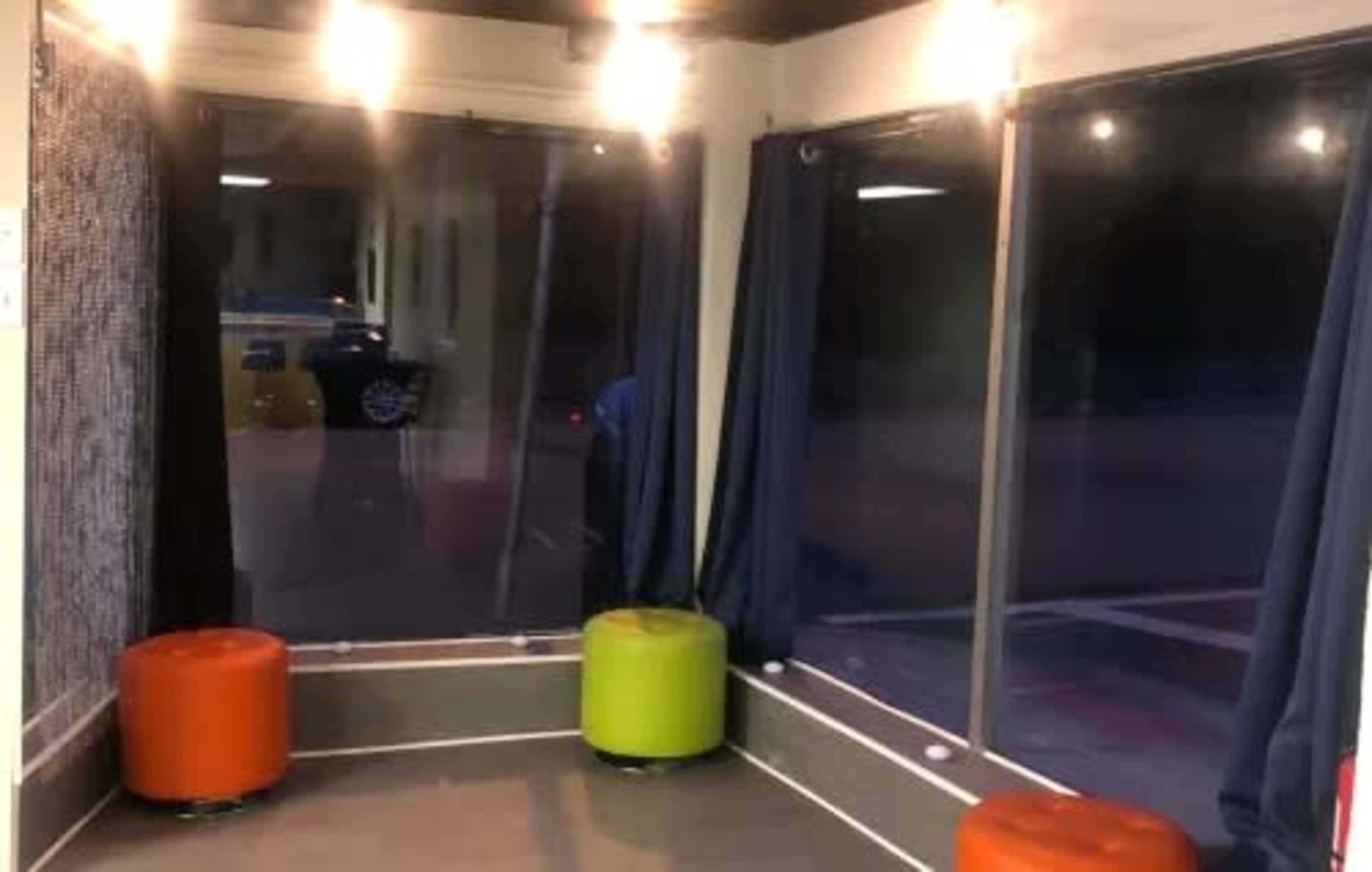 The image shows a corner of a room with large windows, two circular seating stools—one orange and one green—and dark curtains.