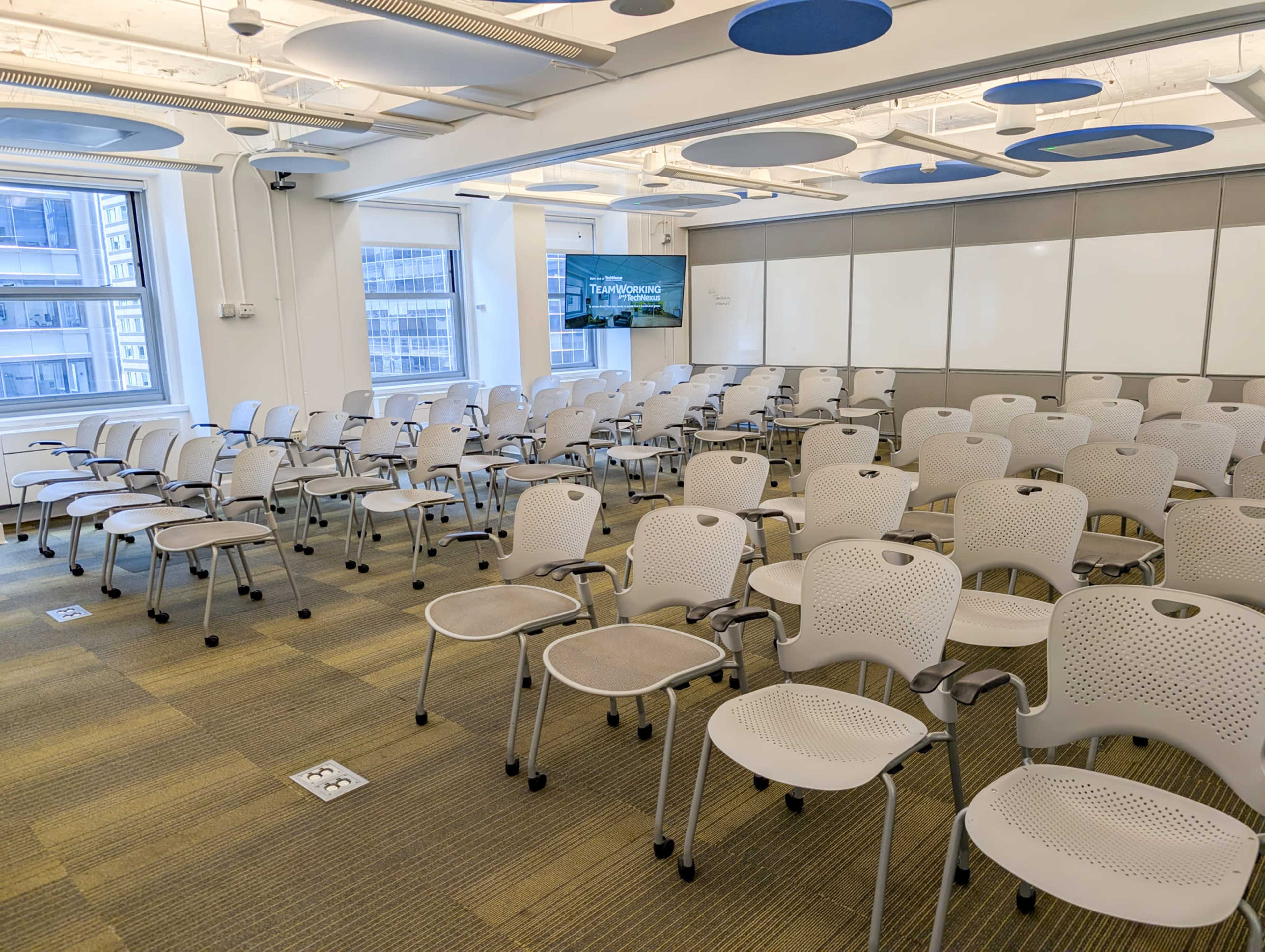 The image shows a conference room arranged with multiple rows of white chairs facing a screen and whiteboards.