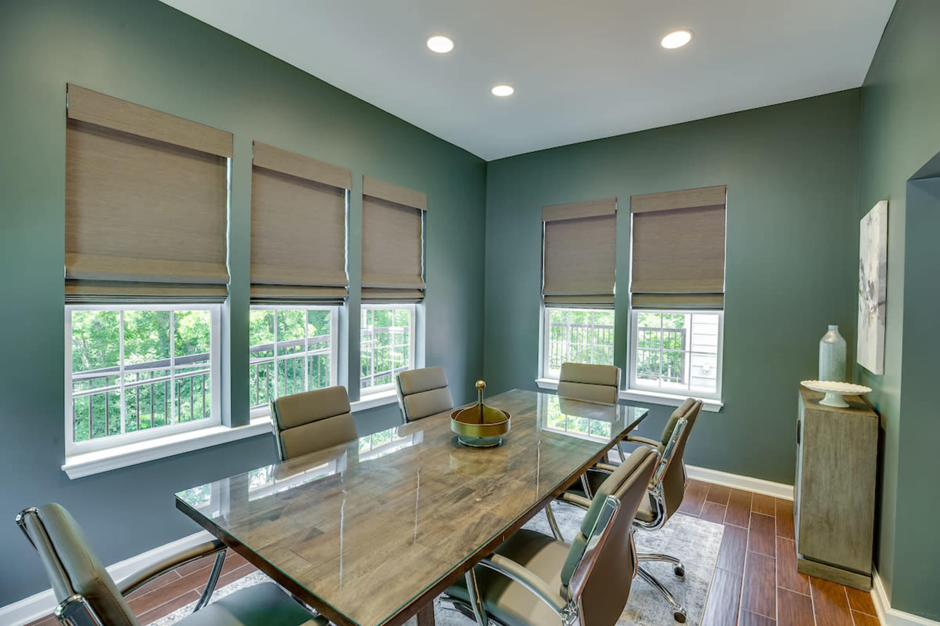 A modern conference room with a glass table surrounded by silver chairs, featuring large windows covered with brown shades.