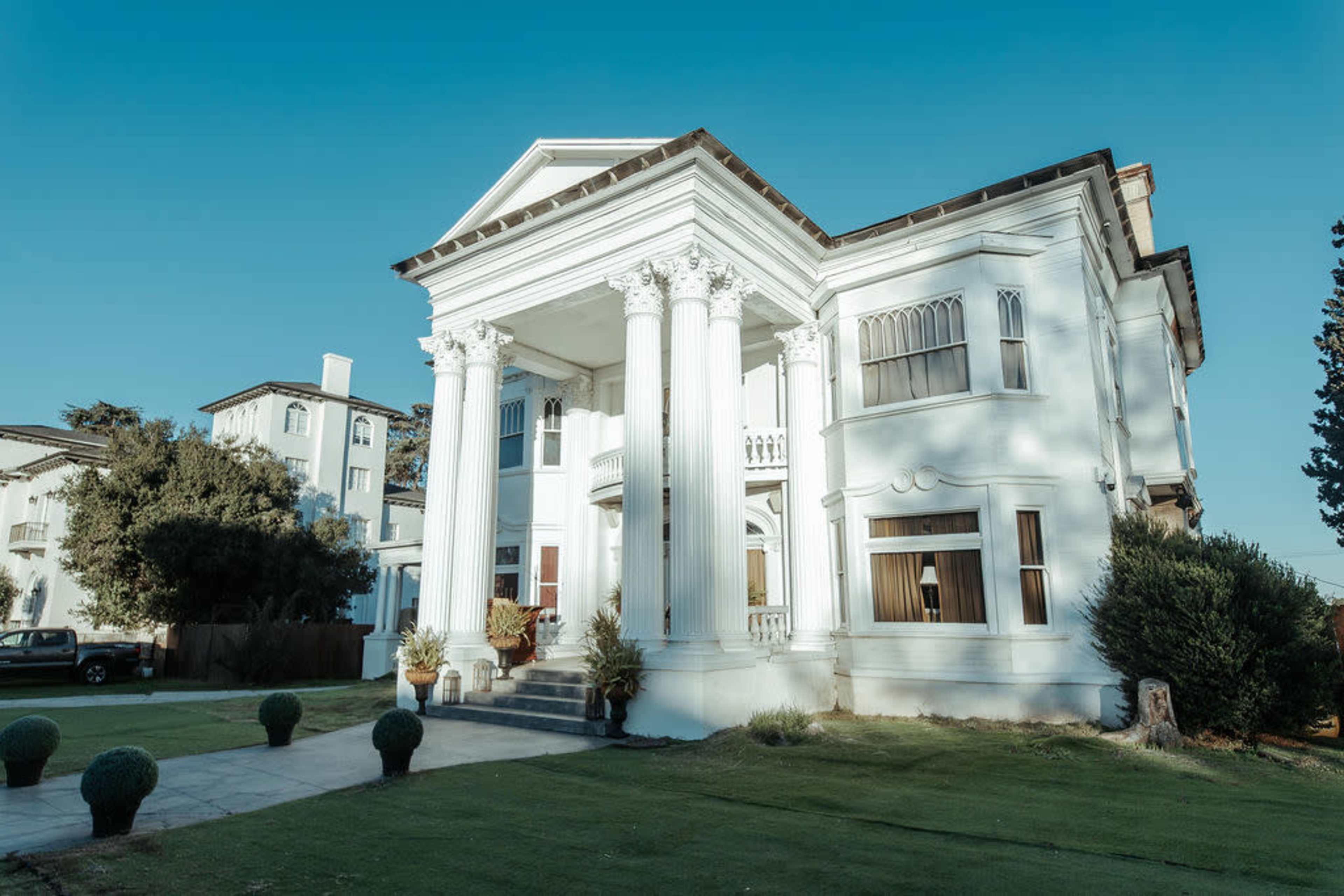 The image shows a large, classical-style mansion with white columns and a landscaped front yard.