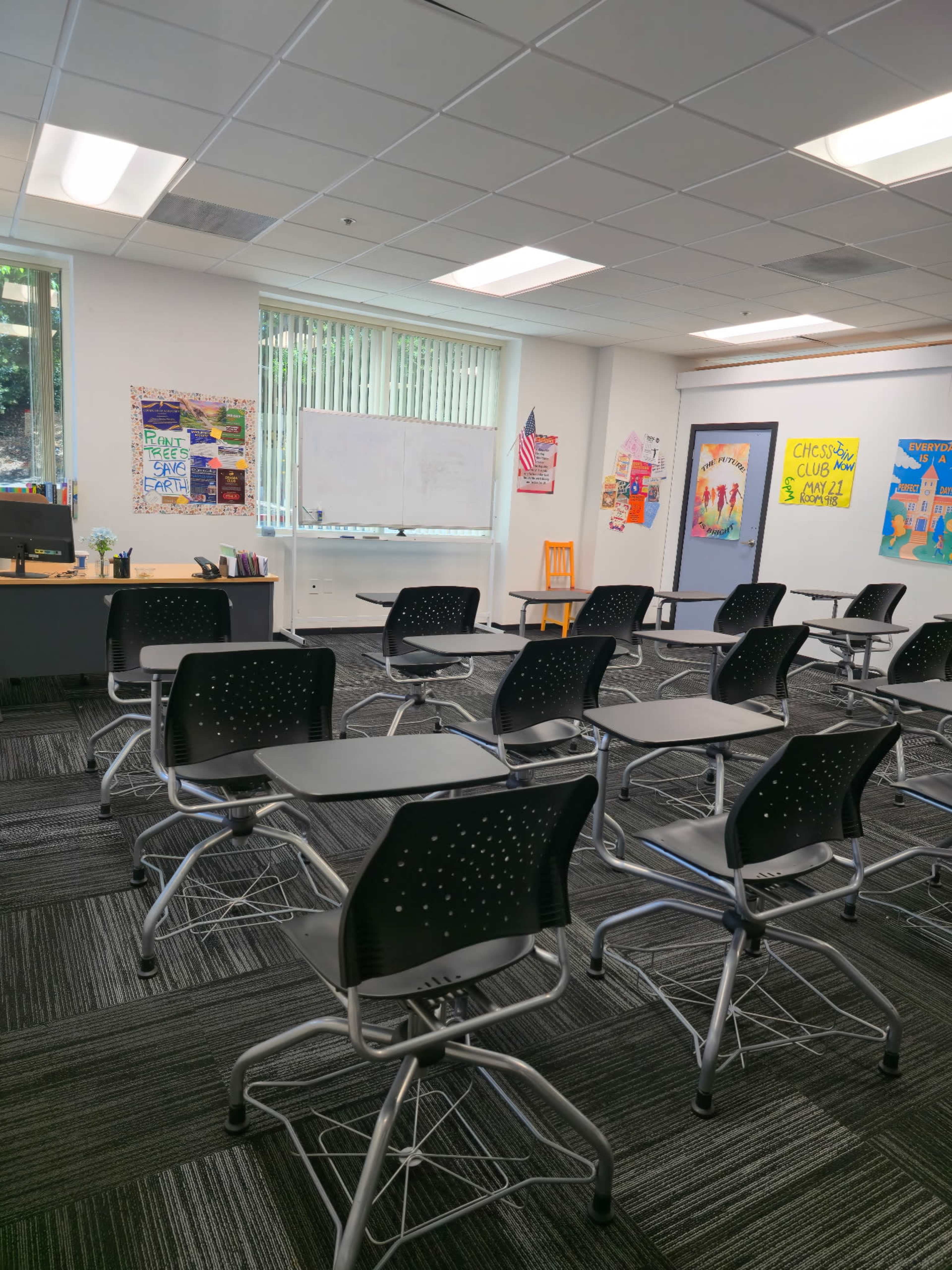 The classroom features desks arranged in rows facing a whiteboard, with colorful posters on the walls and a teacher’s desk in the corner.