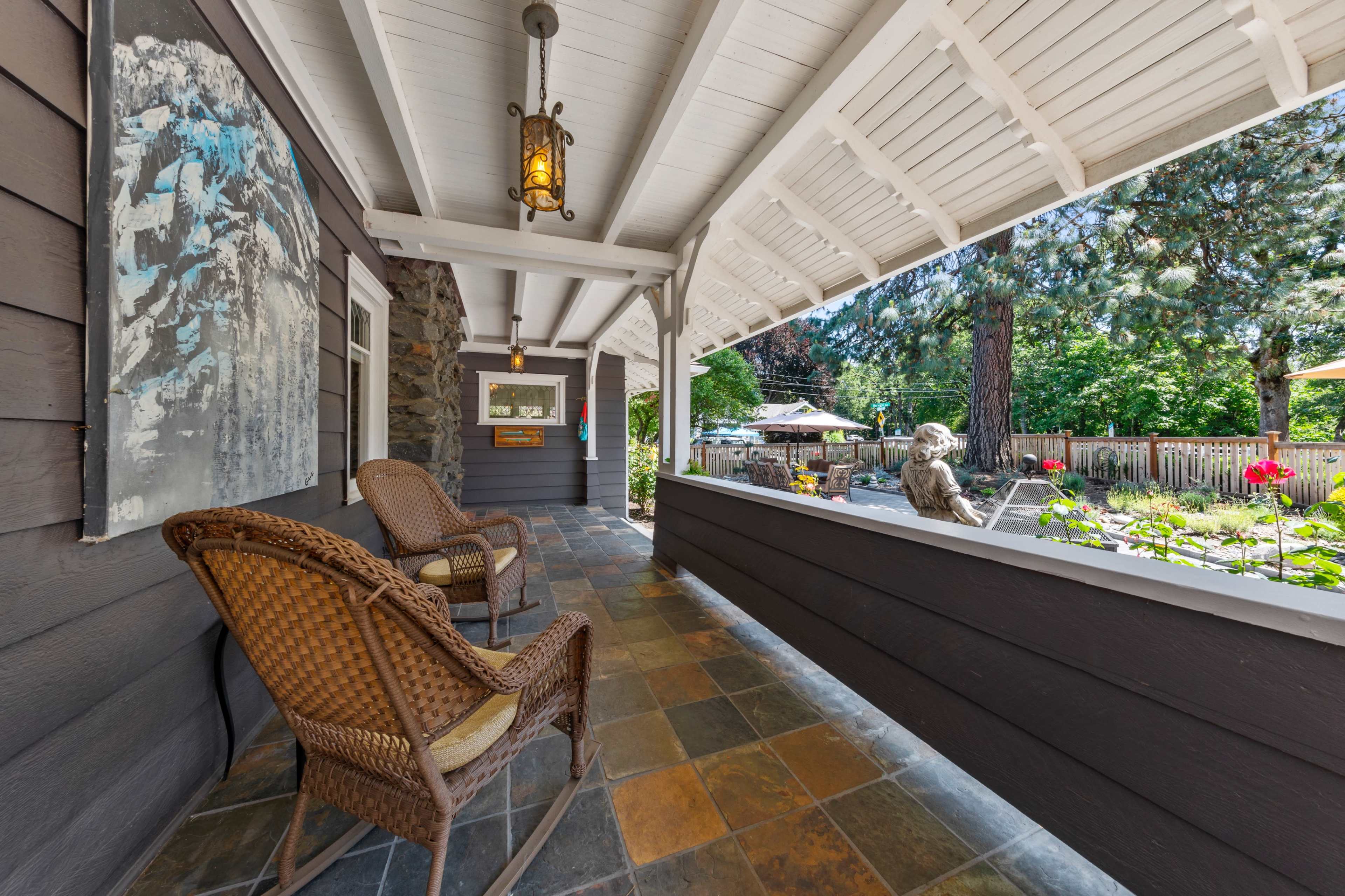The image shows a covered porch with two woven chairs, a stone pathway, and a view of a well-manicured garden beyond.