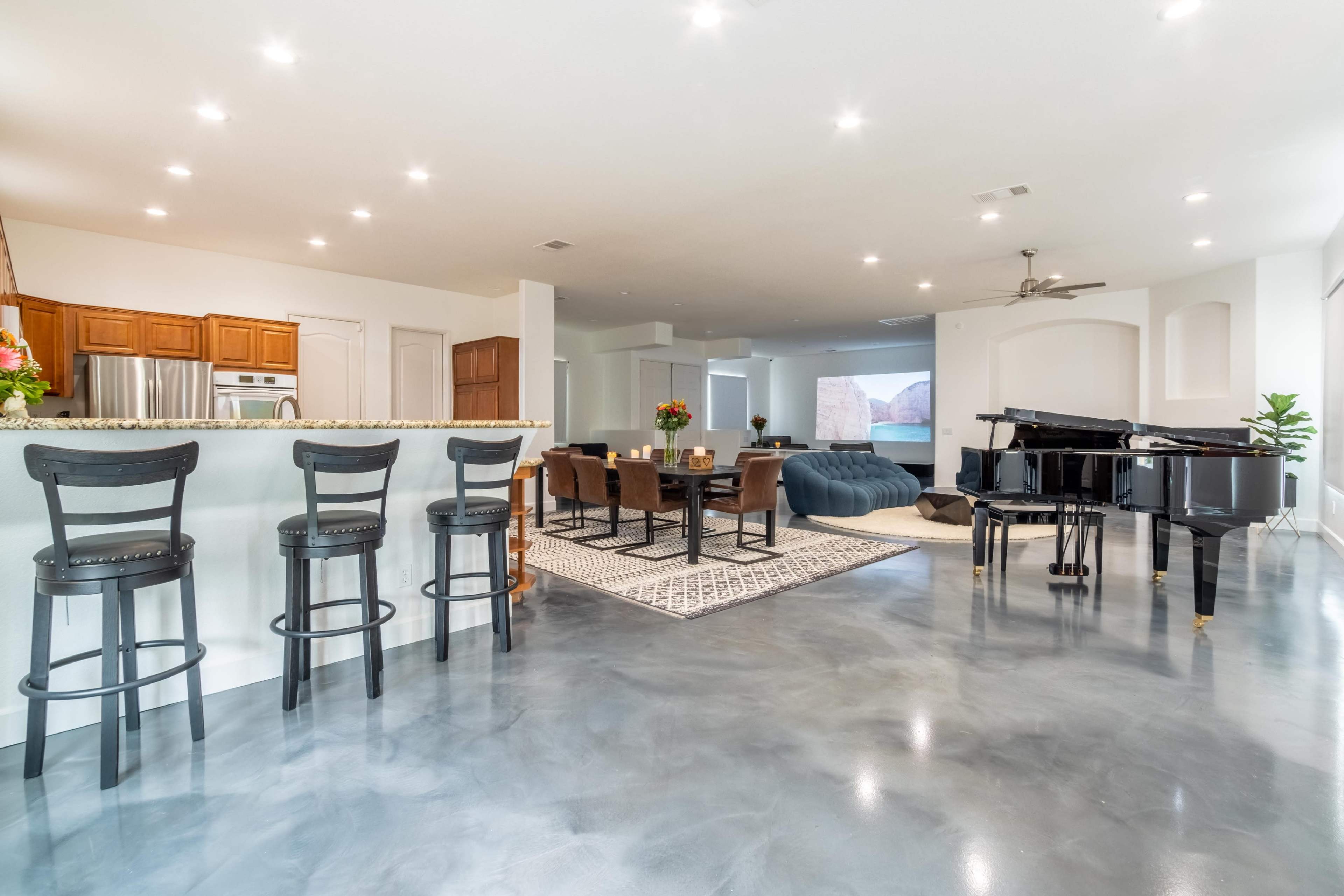 A modern open-concept living space featuring a kitchen with bar stools, a dining area, and a piano, all illuminated by recessed lighting.