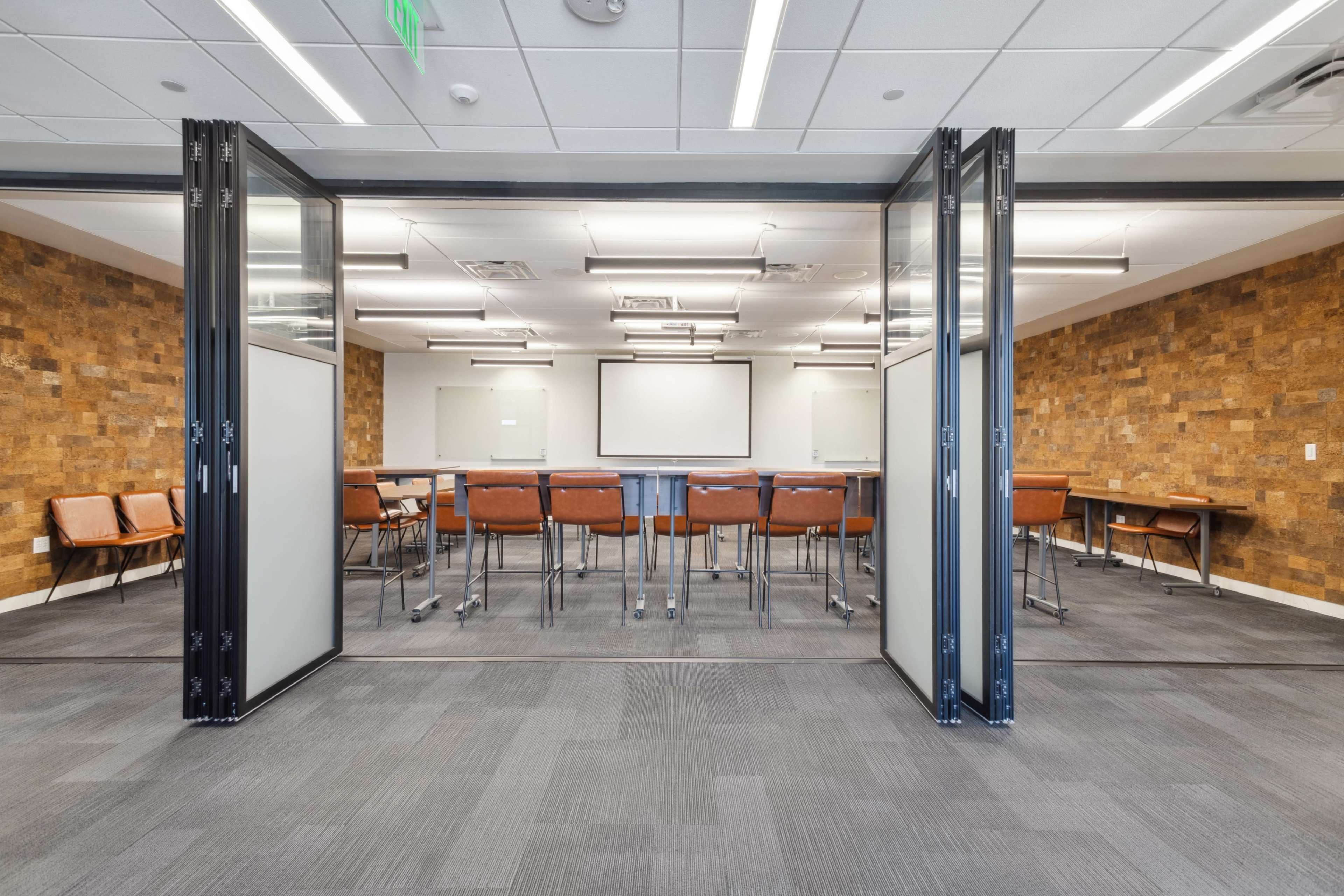 A conference room features a long table surrounded by chairs, with a presentation screen visible on a wall and a textured stone feature wall in the background.
