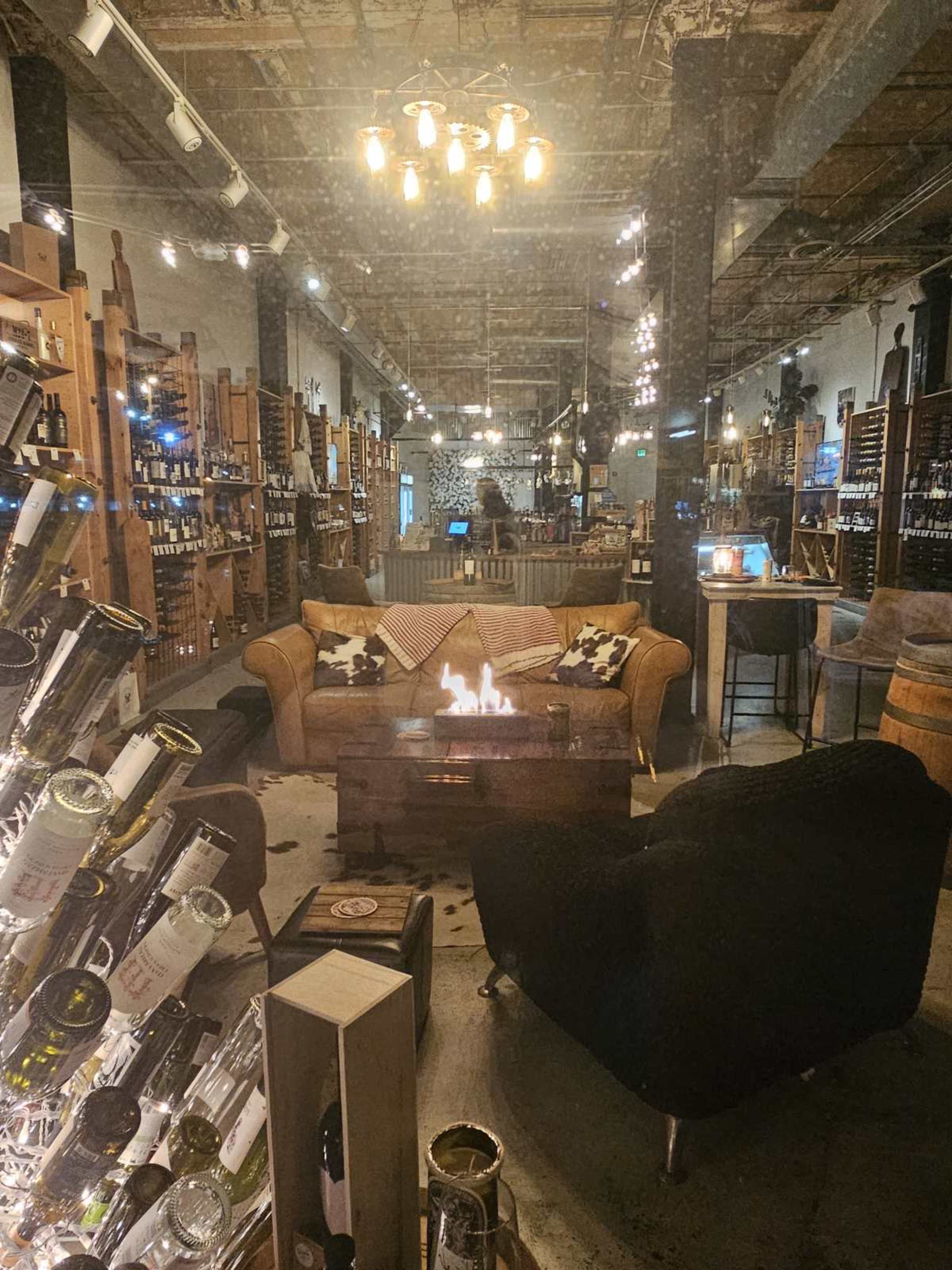 The image shows a cozy seating area with a small fireplace, surrounded by shelves filled with bottles in a wine shop.