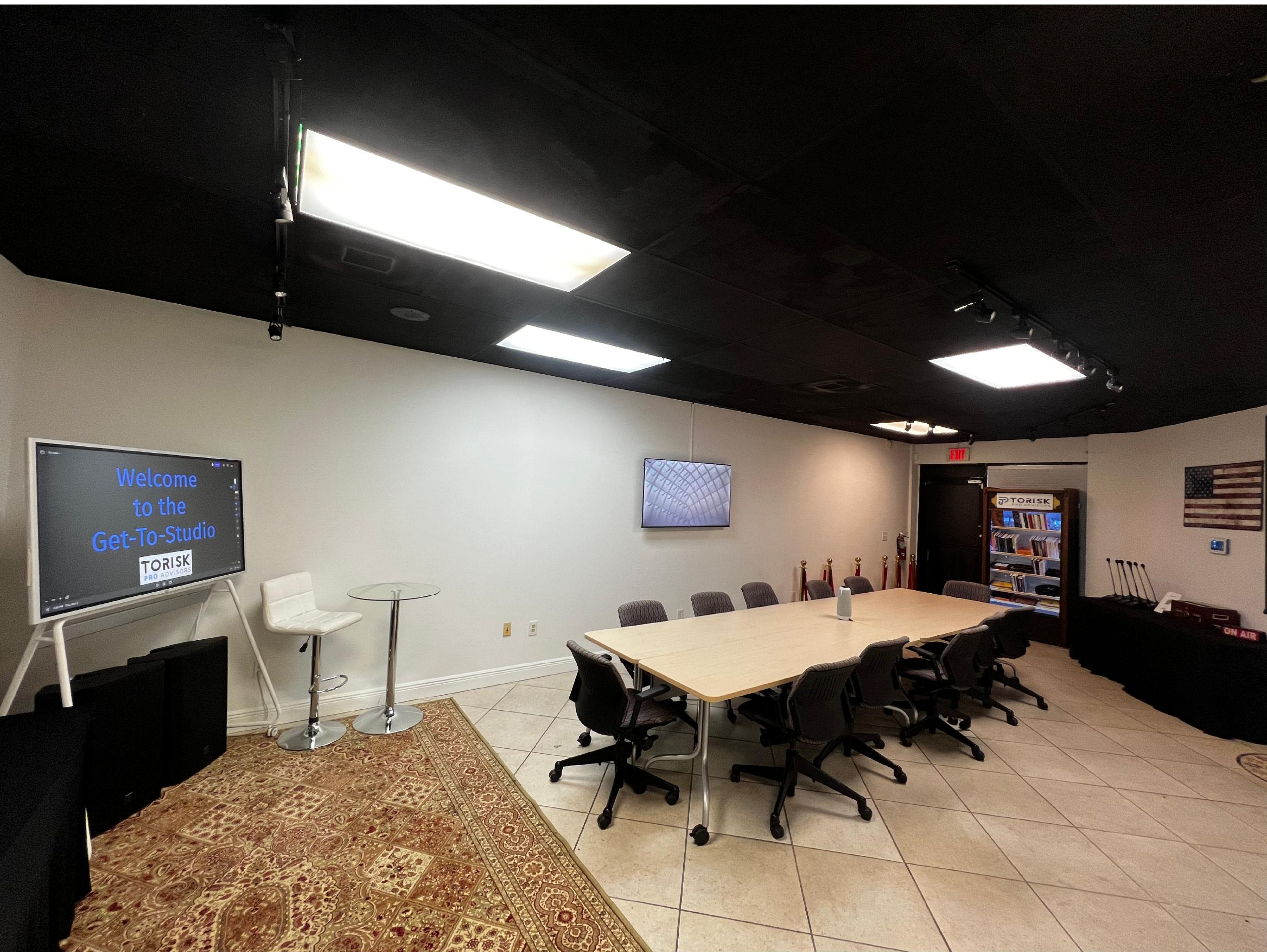 The image shows a conference room with a large rectangular table surrounded by chairs, a screen displaying a welcome message, and additional wall-mounted displays.