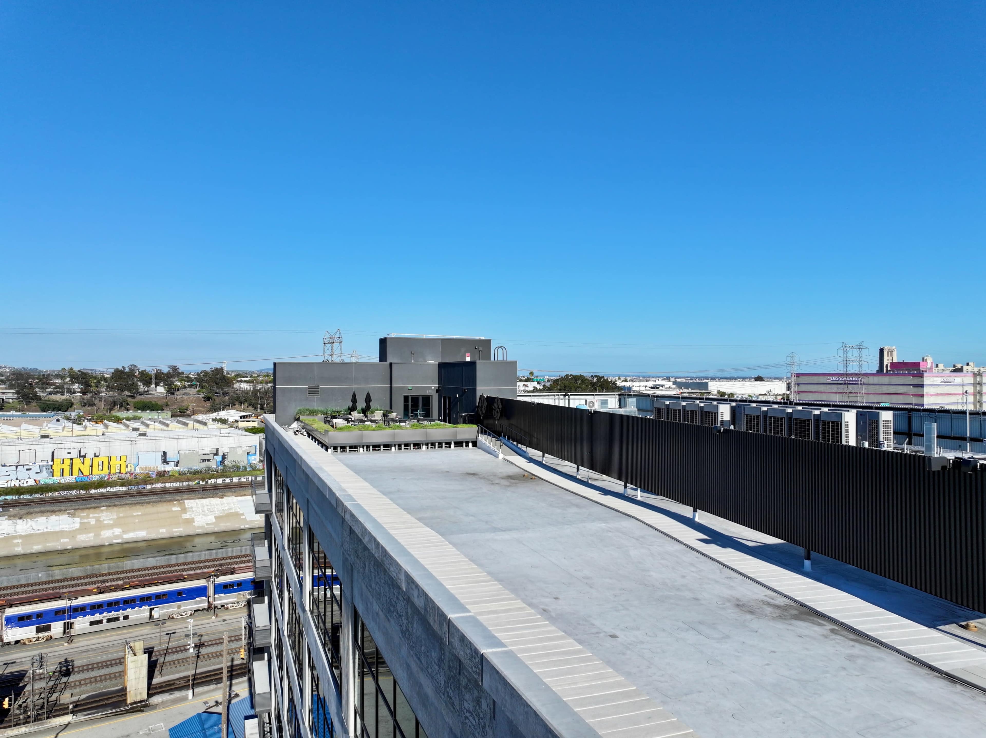A rooftop terrace with green plants overlooks a railway line and industrial buildings under a clear blue sky.