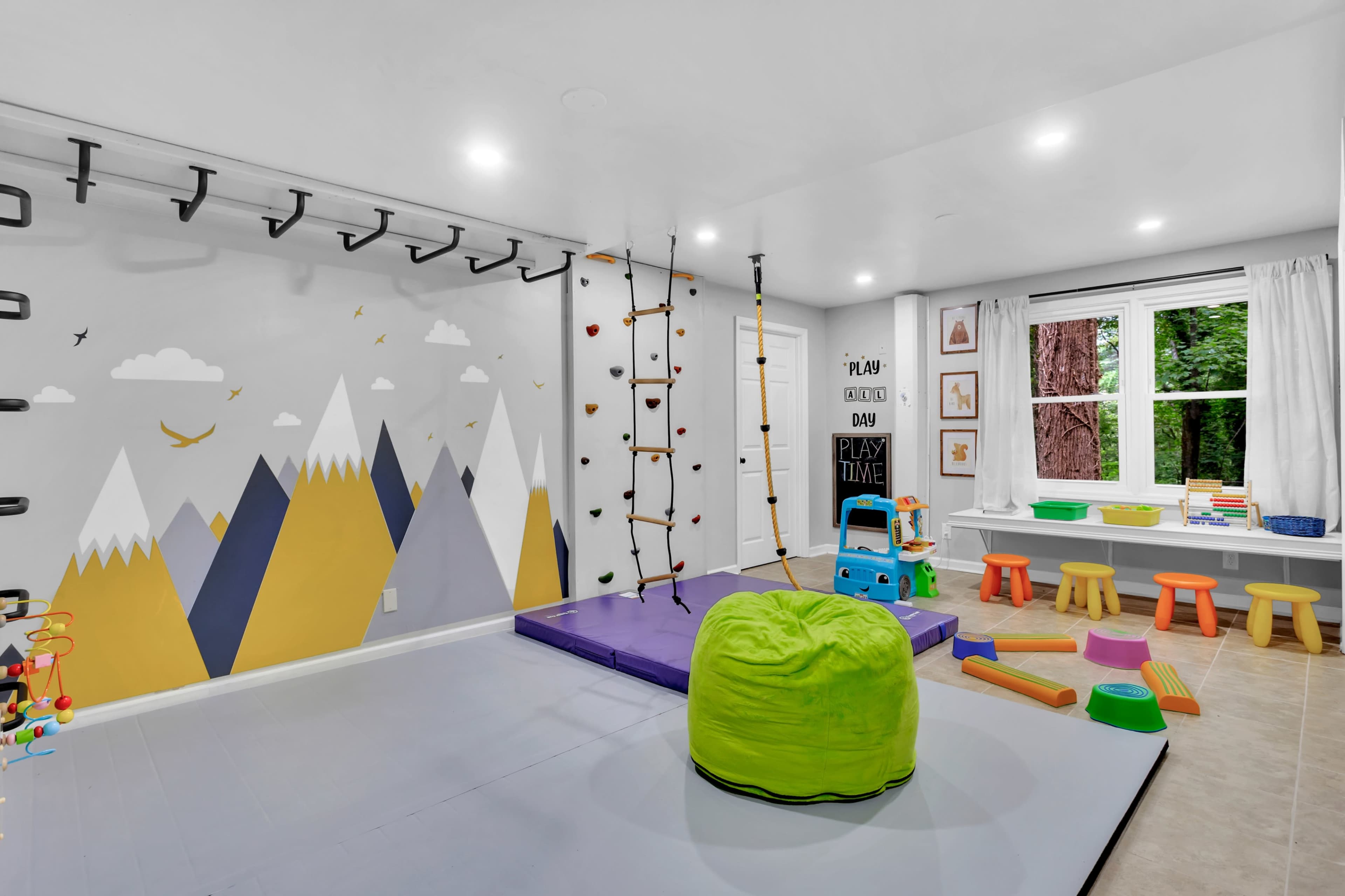 The image shows a brightly lit children's playroom featuring a climbing wall, colorful seating, and various toys, with mountain-themed wall art and a large window overlooking greenery.
