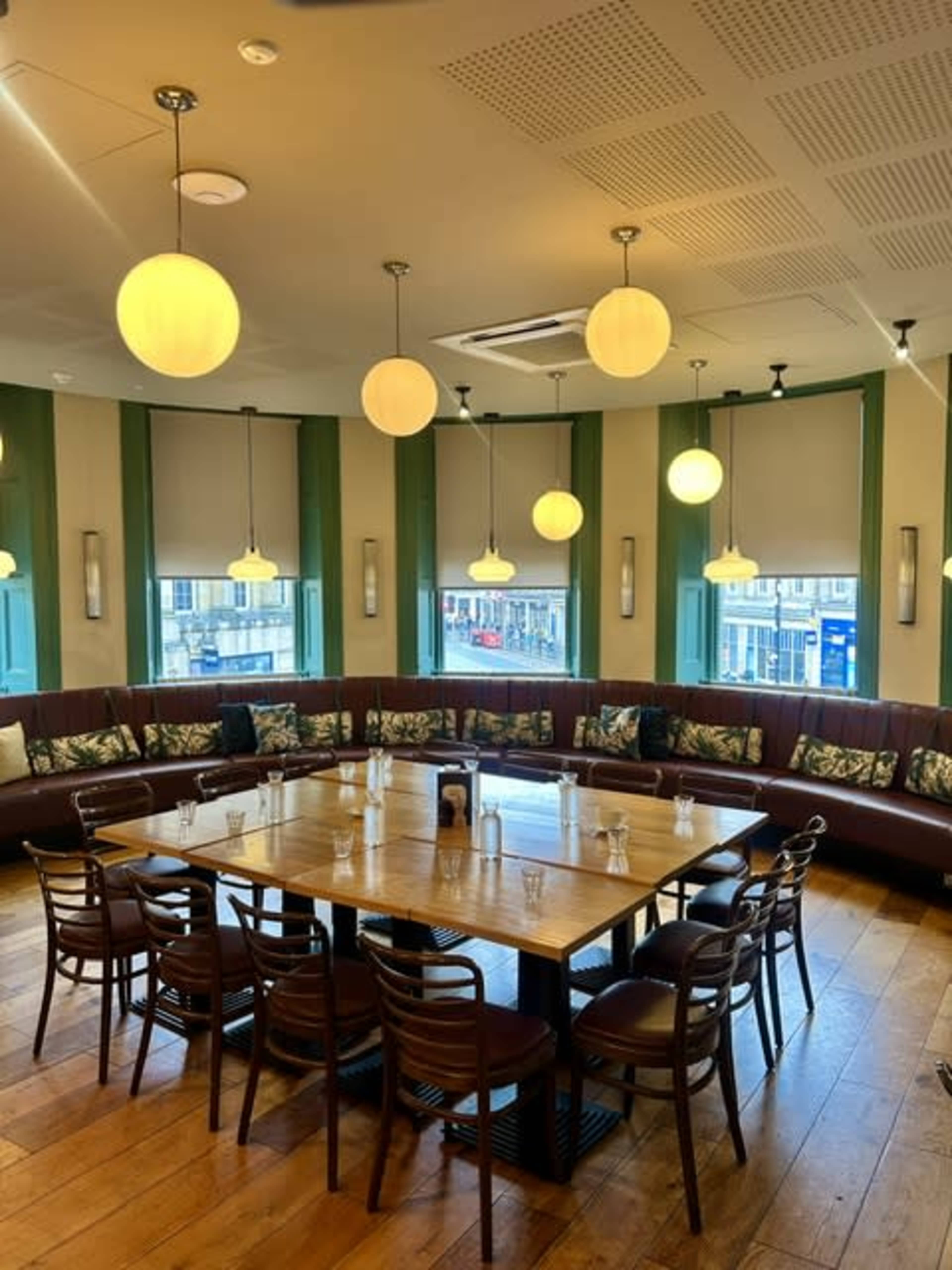 The image shows a dining area with a large, round wooden table surrounded by chairs in a corner with green accents and overhead pendant lights.