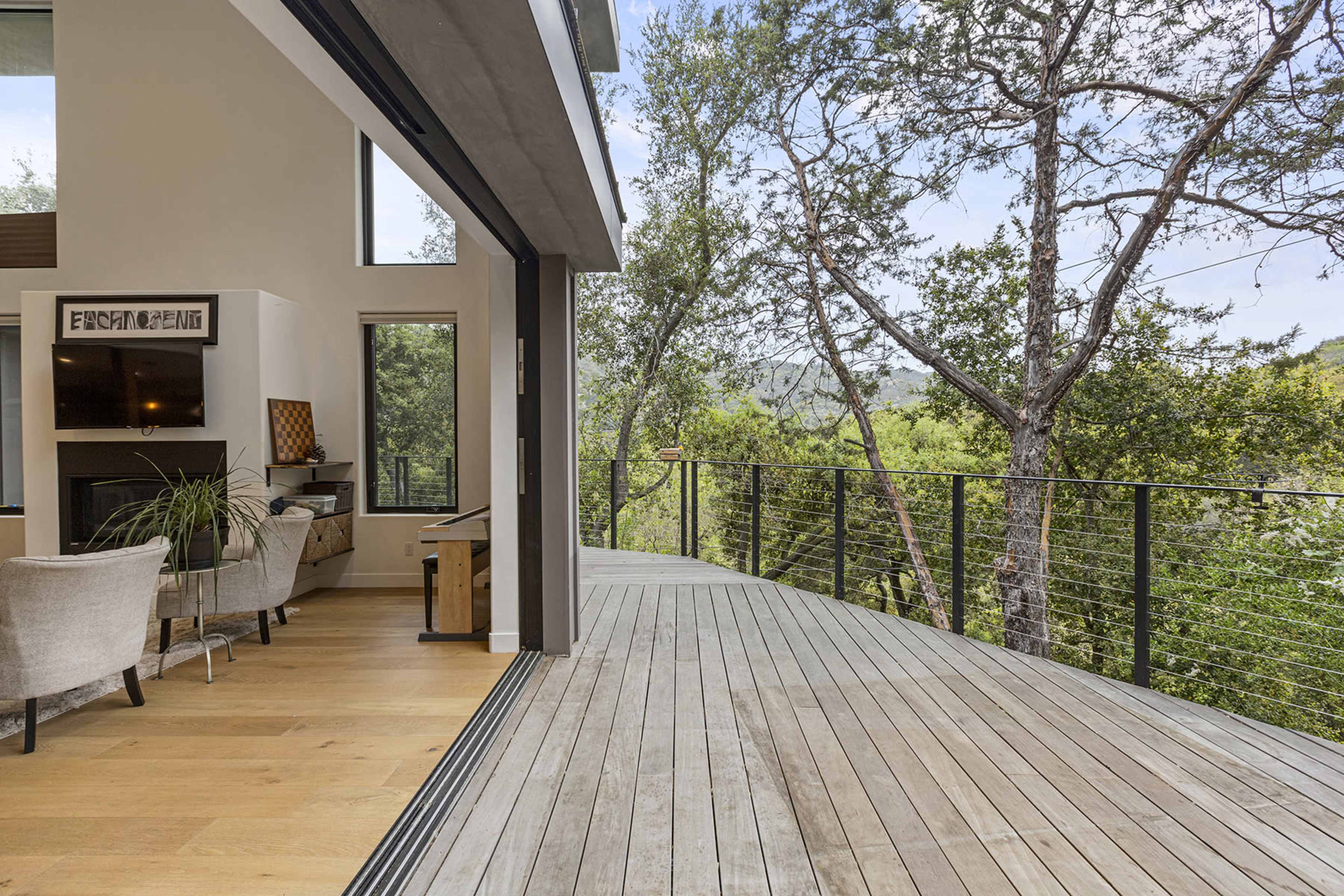 A modern interior space with large windows opens onto a wooden deck surrounded by trees and greenery.