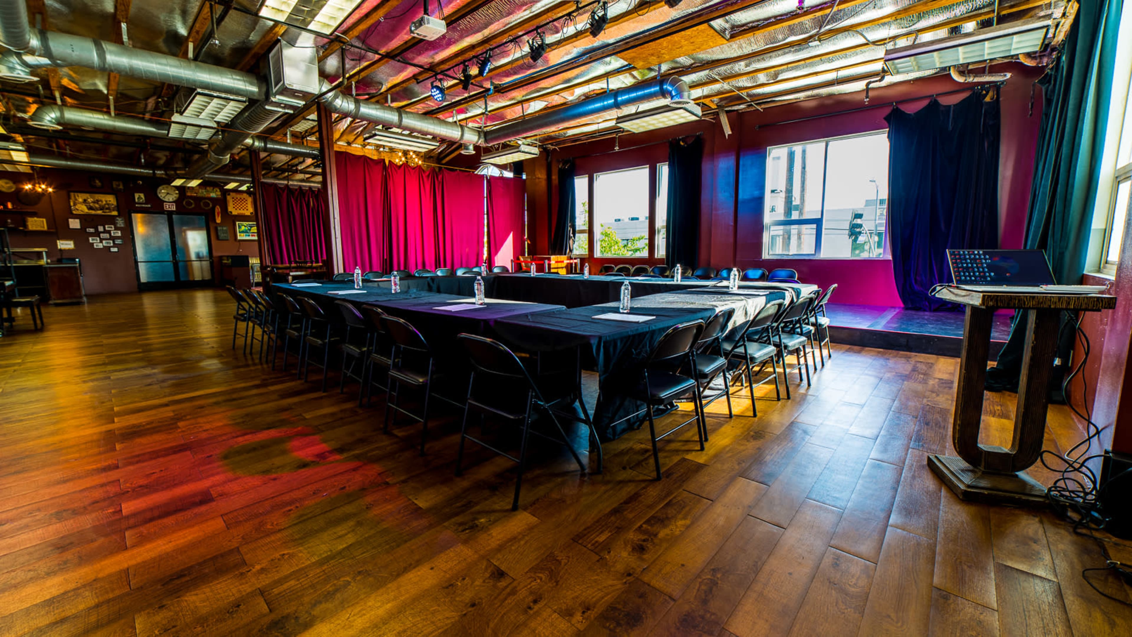 A conference room features a long rectangular table set with chairs, surrounded by red and purple drapes, with large windows allowing natural light to illuminate the wooden floor.