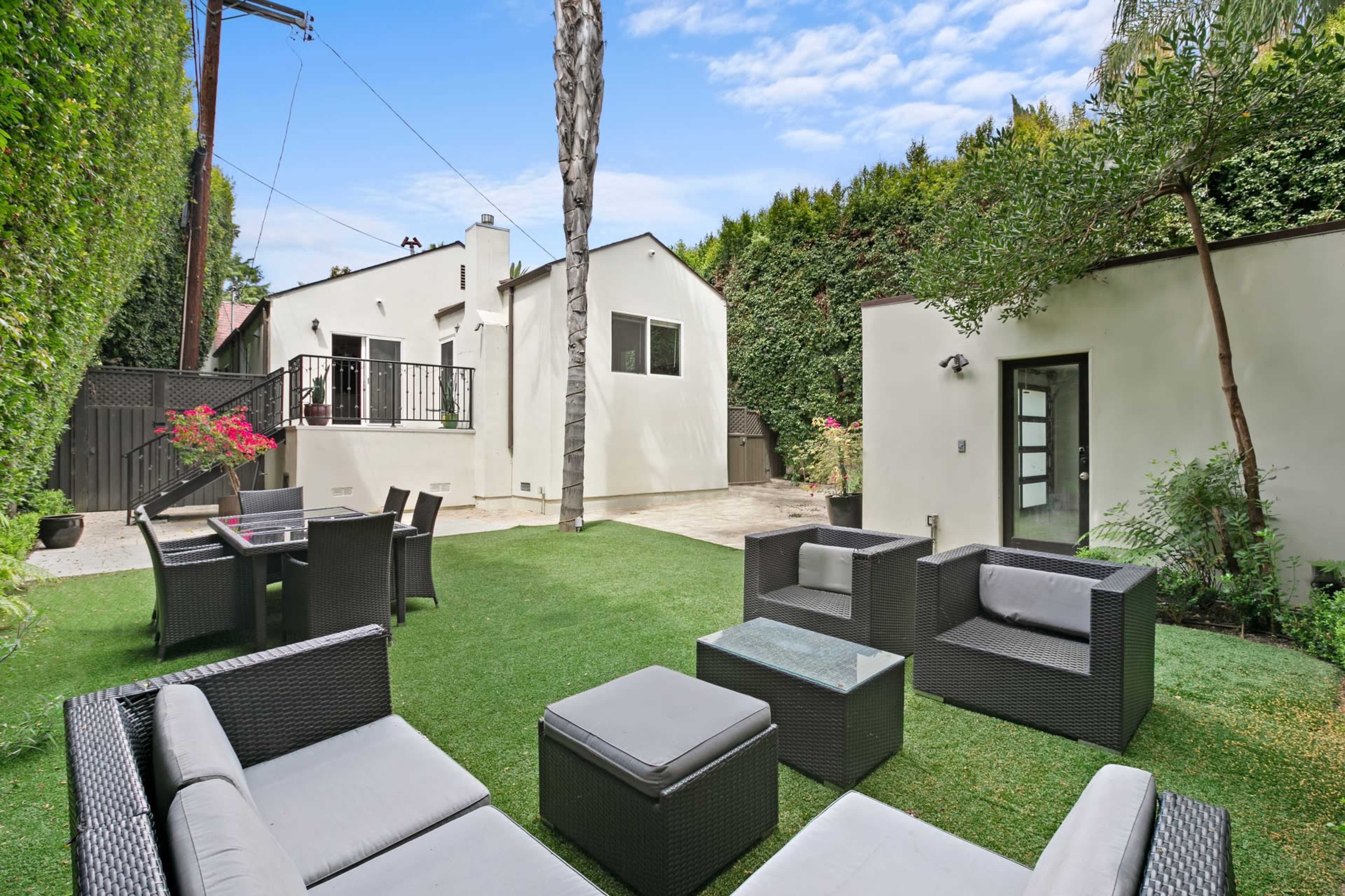 The image shows a backyard patio with modern seating arrangements, a small dining area, and greenery surrounding two white houses.