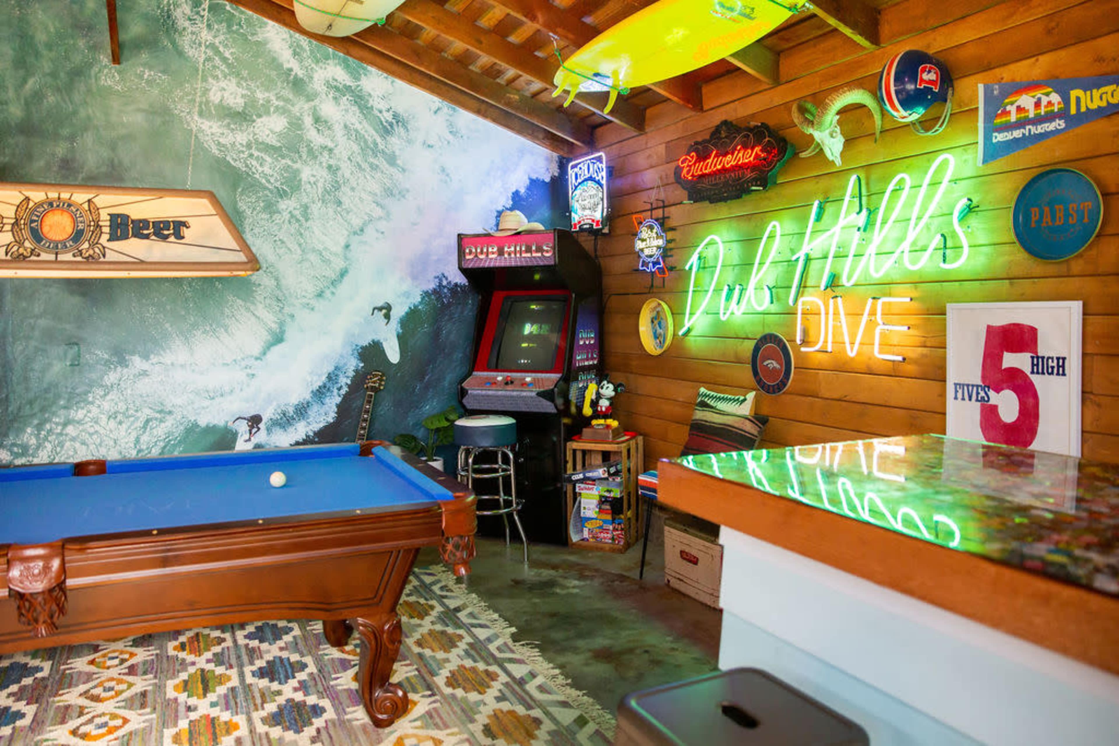 A room with a blue pool table, a retro arcade game, neon signs, and surfboard decor against a mural of waves.