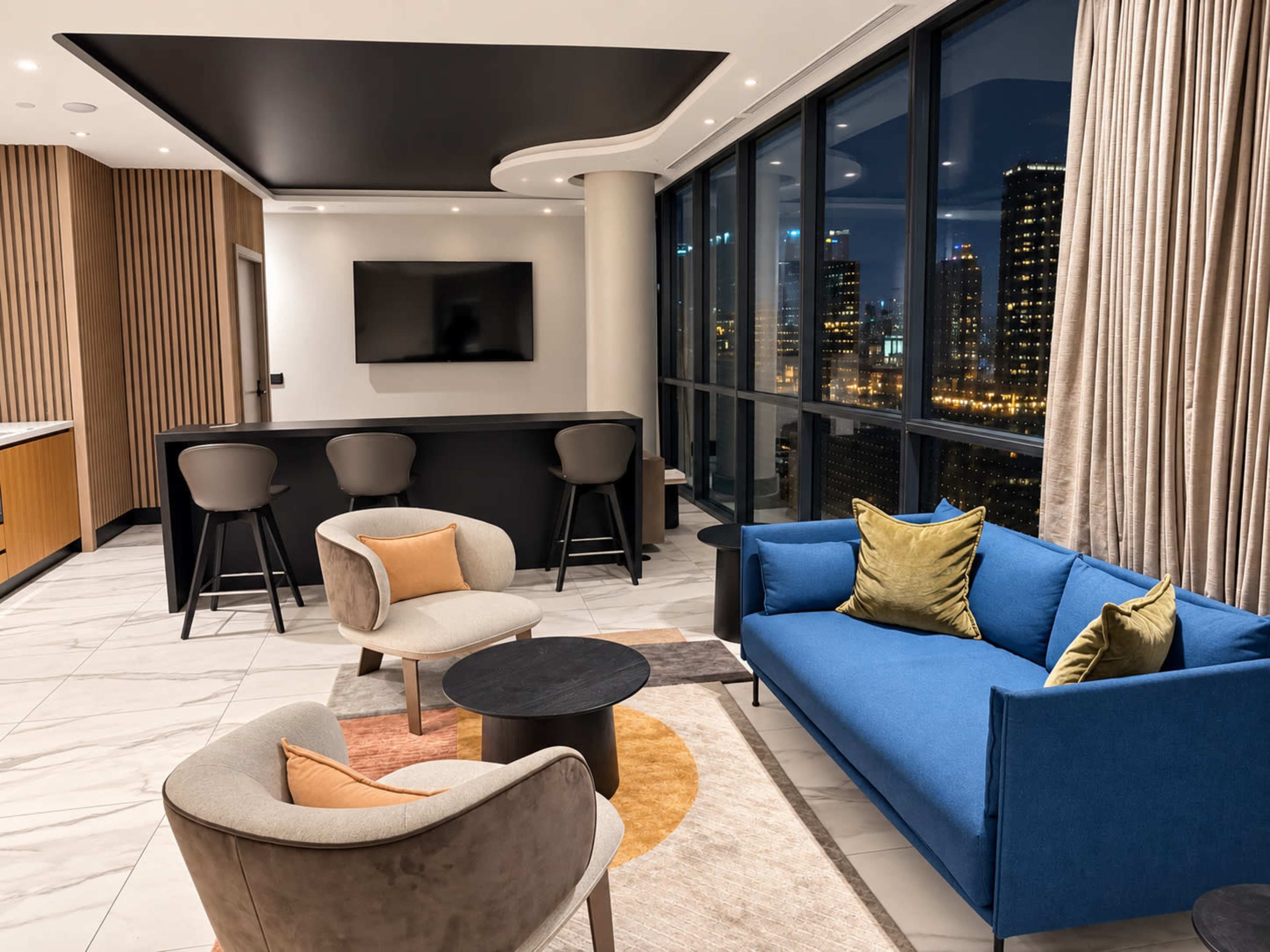 A modern living room features a blue sofa, two armchairs, a round coffee table, and a bar area with high stools, all set against large windows showcasing a city skyline.