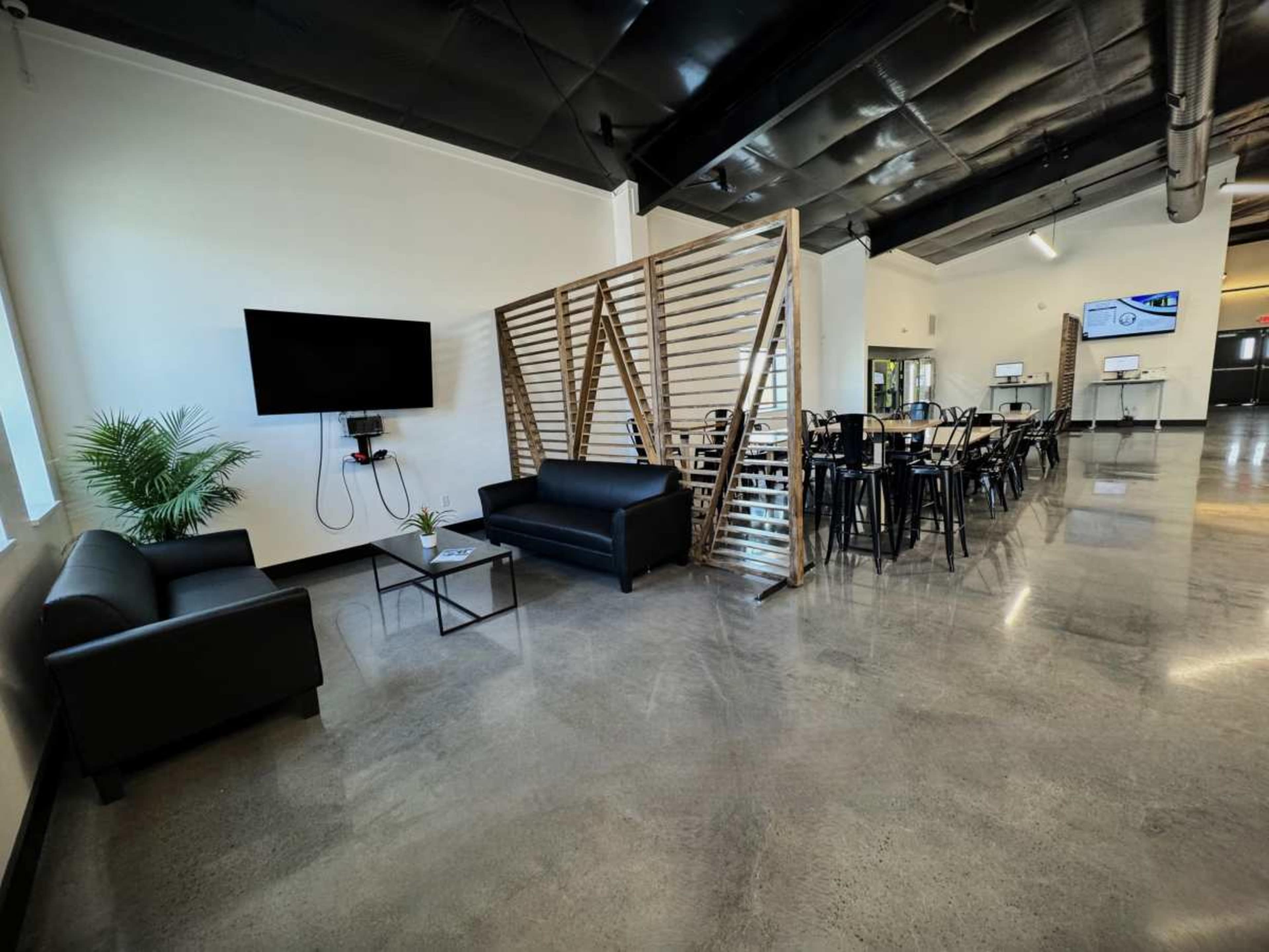A modern interior space featuring a seating area with black sofas, a television mounted on the wall, a plant, and a dining area with a long table and chairs.