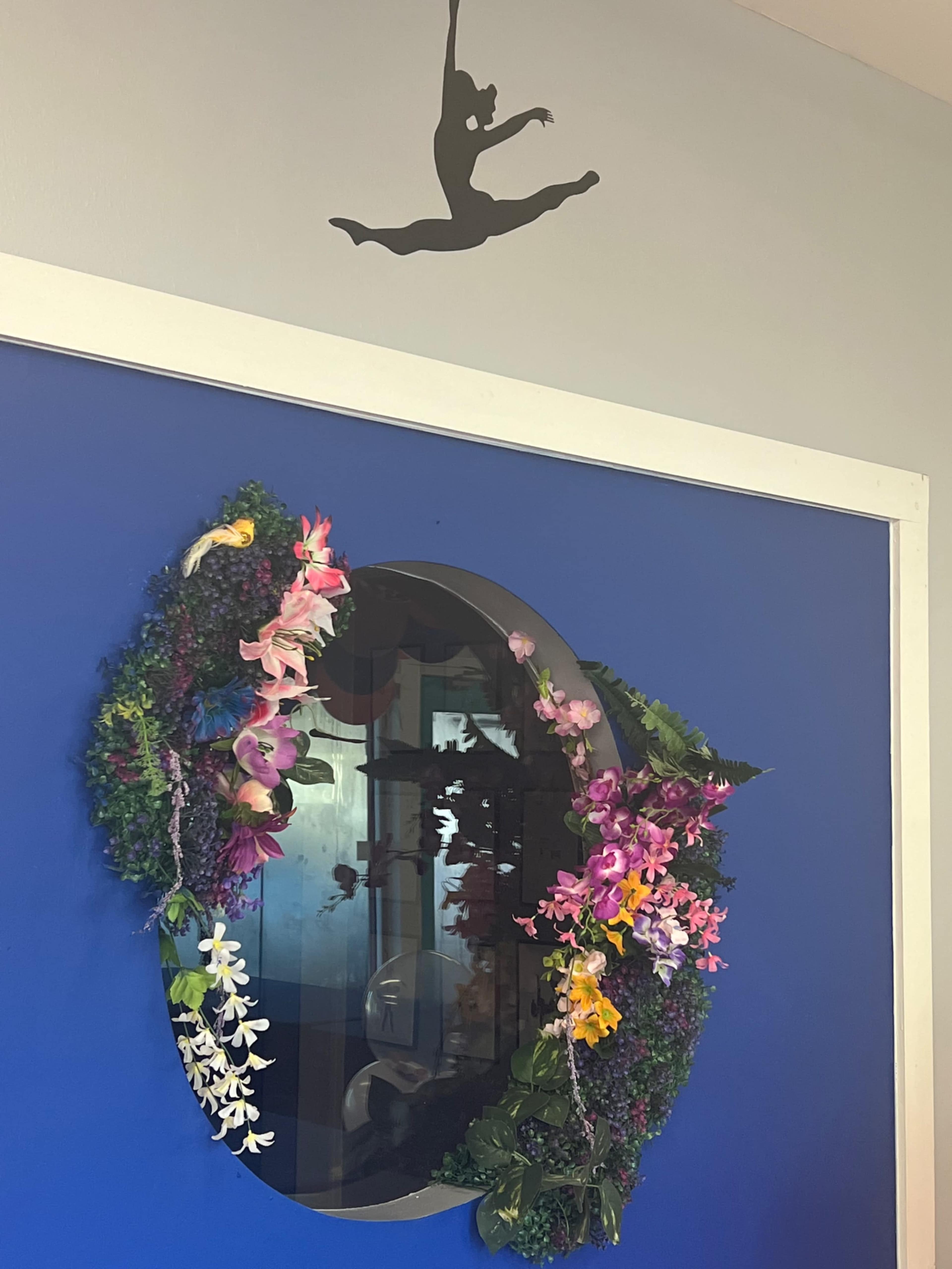A round mirror surrounded by a floral wreath is mounted on a blue wall beneath a silhouette of a dancer.