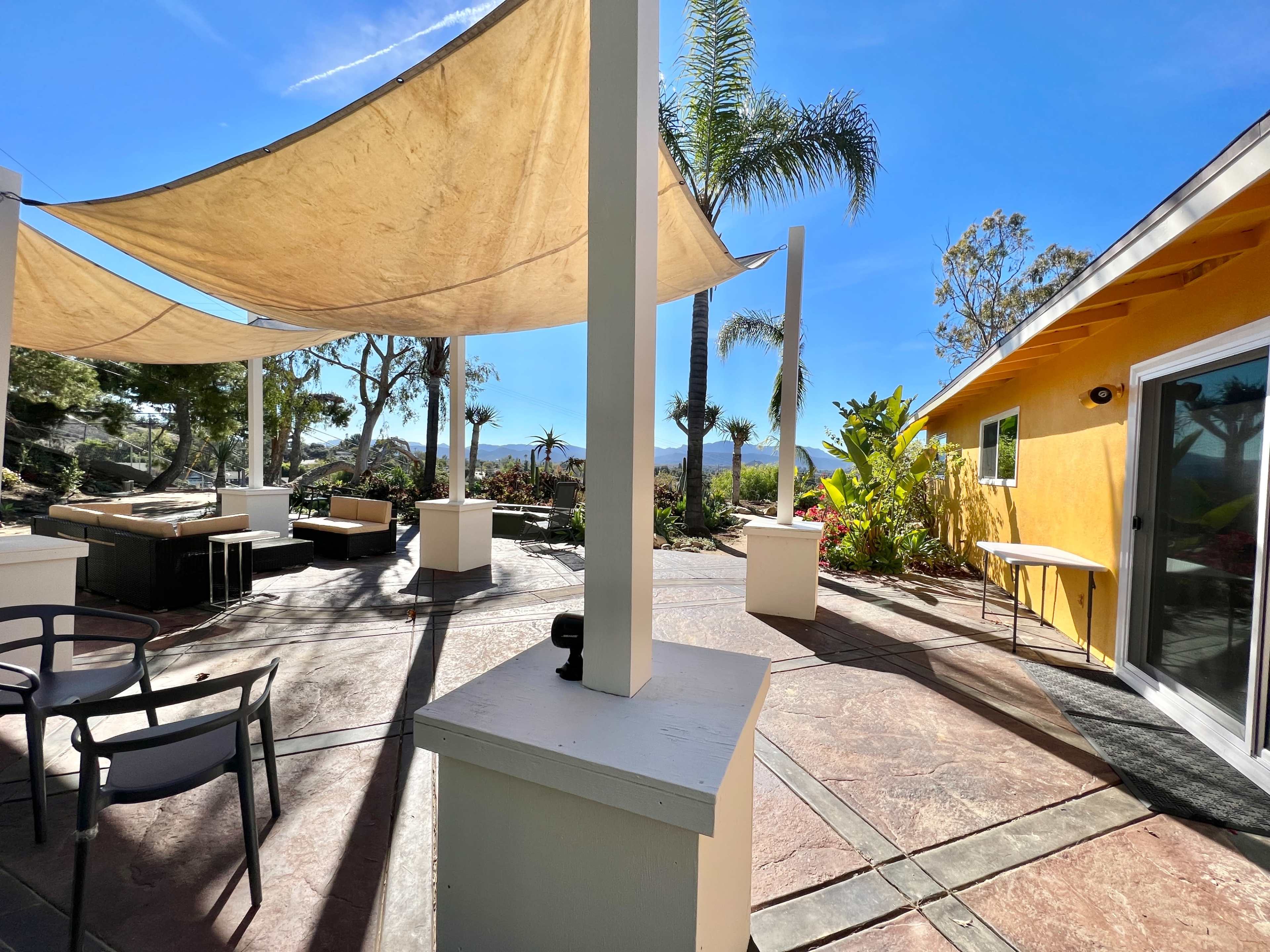 An outdoor patio area with a yellow building, shaded seating under awnings, and palm trees in the background.