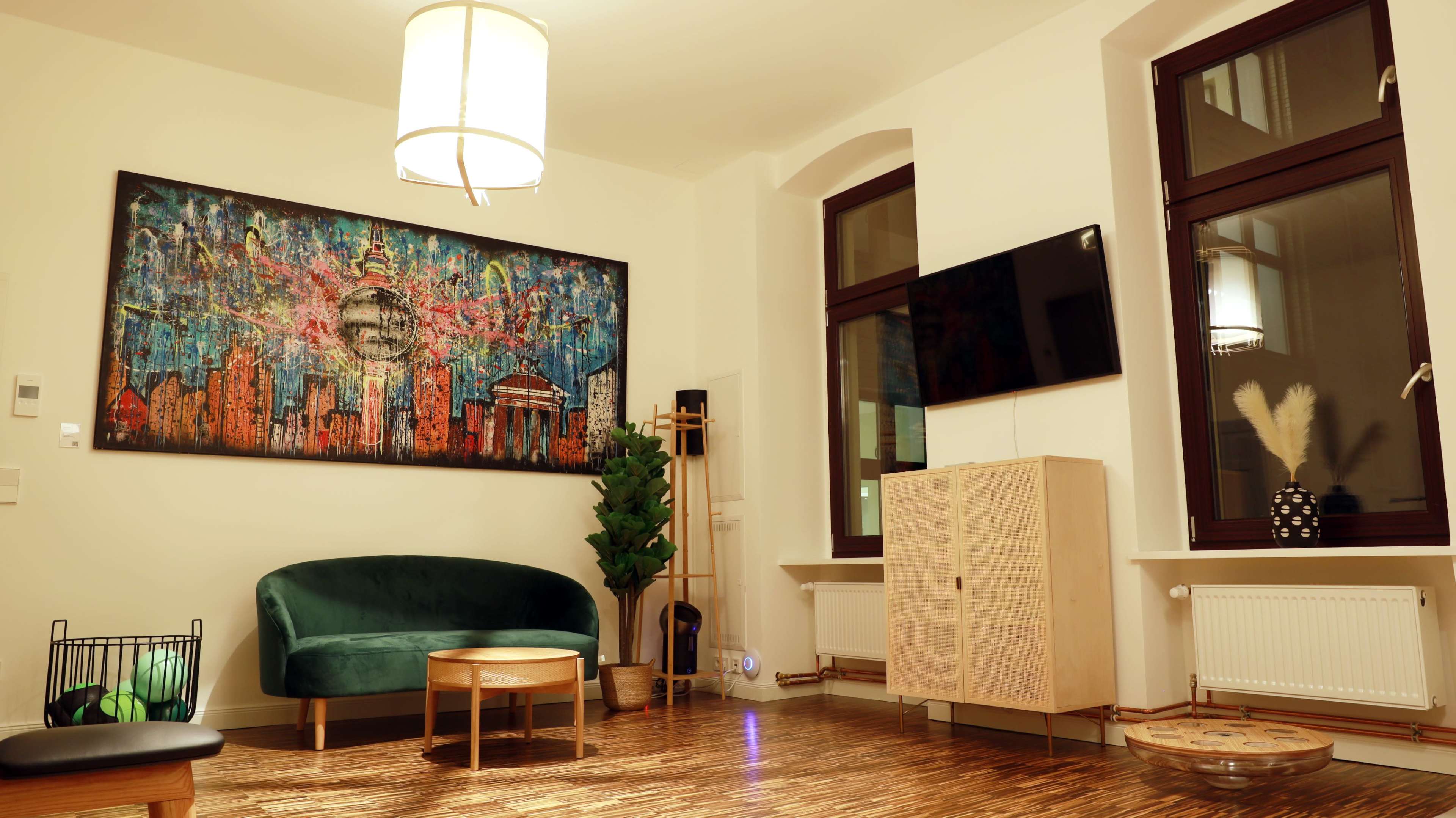 The image shows a modern living room with a green sofa, a wooden coffee table, a large colorful artwork on the wall, and a flat-screen TV mounted opposite the seating area.