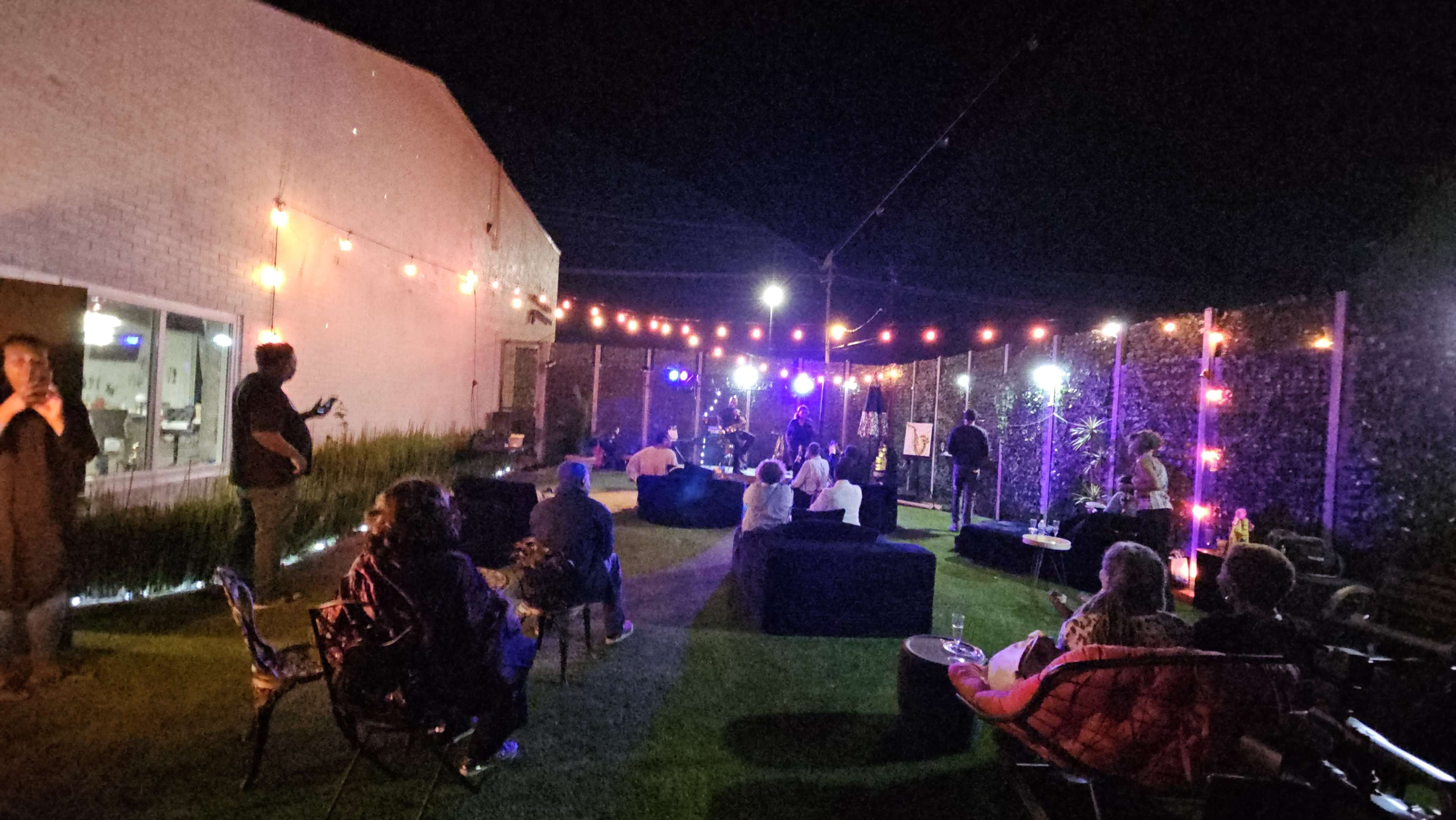 The scene depicts an outdoor gathering at night with people seated in casual seating arrangements, illuminated by string lights and colorful spotlights.