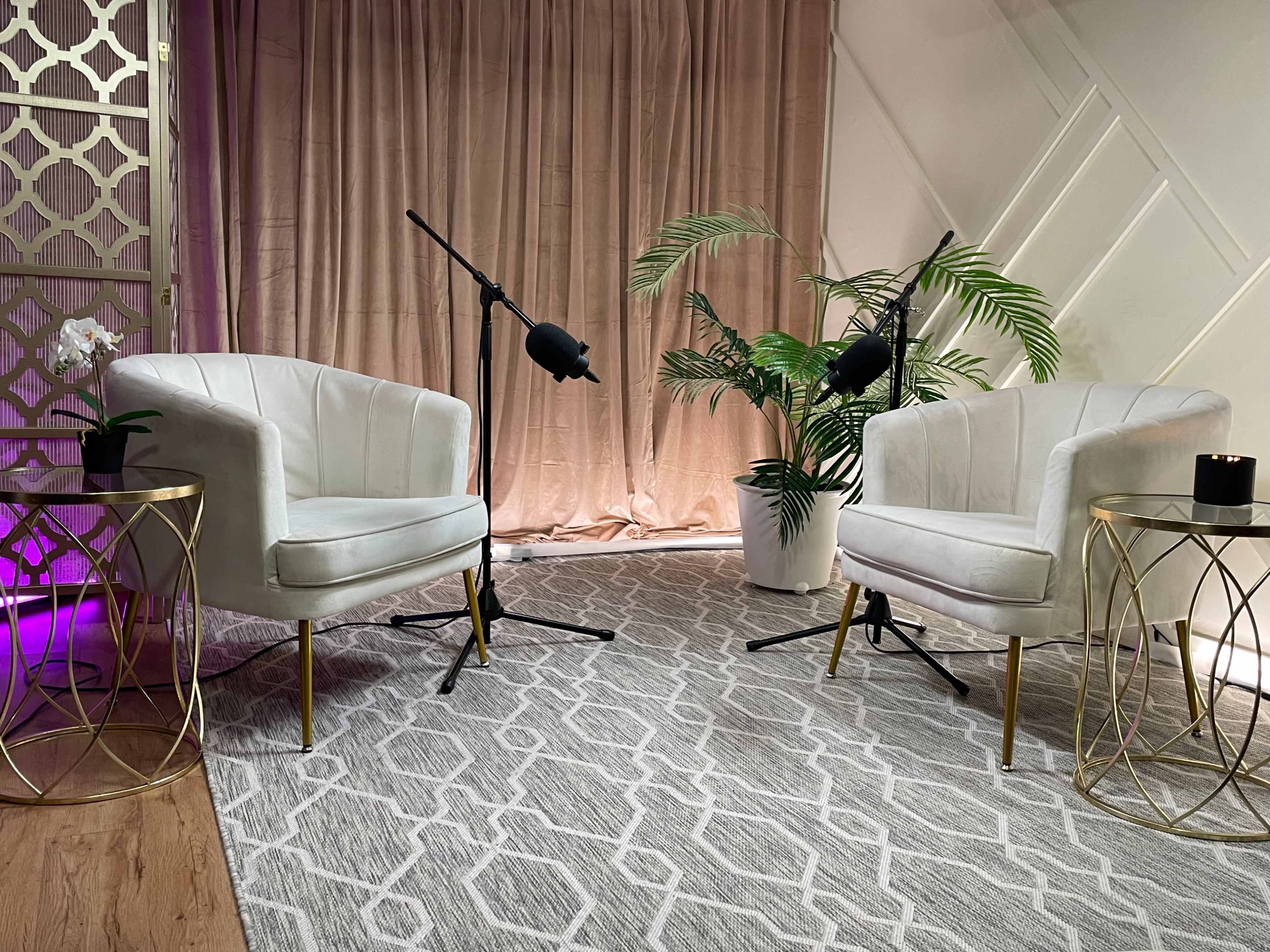The image depicts a modern setting with two white chairs, each accompanied by a microphone stand, placed on a patterned rug in front of a backdrop of curtains and a potted plant.
