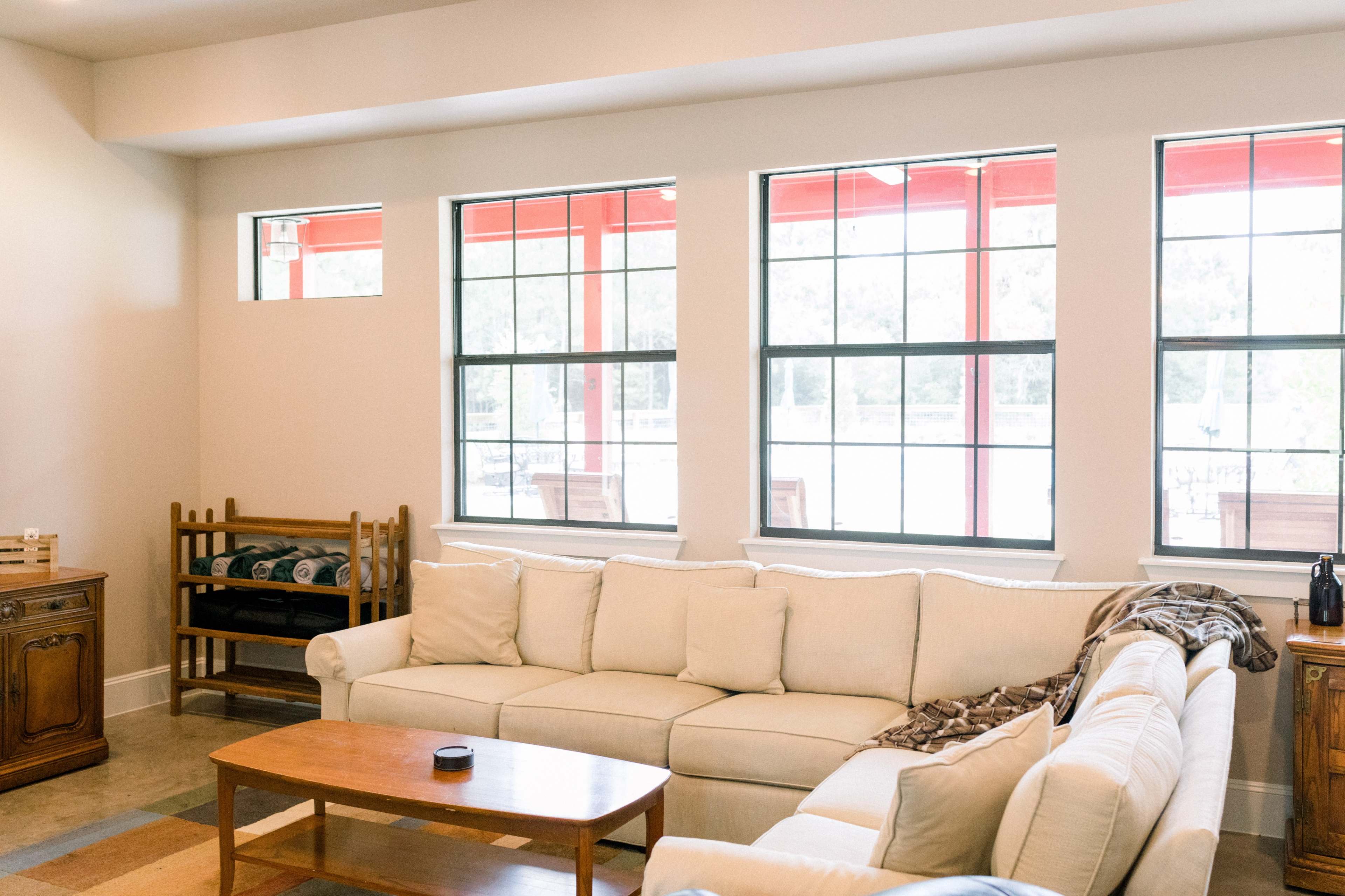 The image shows a bright living room featuring a beige sectional sofa, a wooden coffee table, and large windows with red frames.