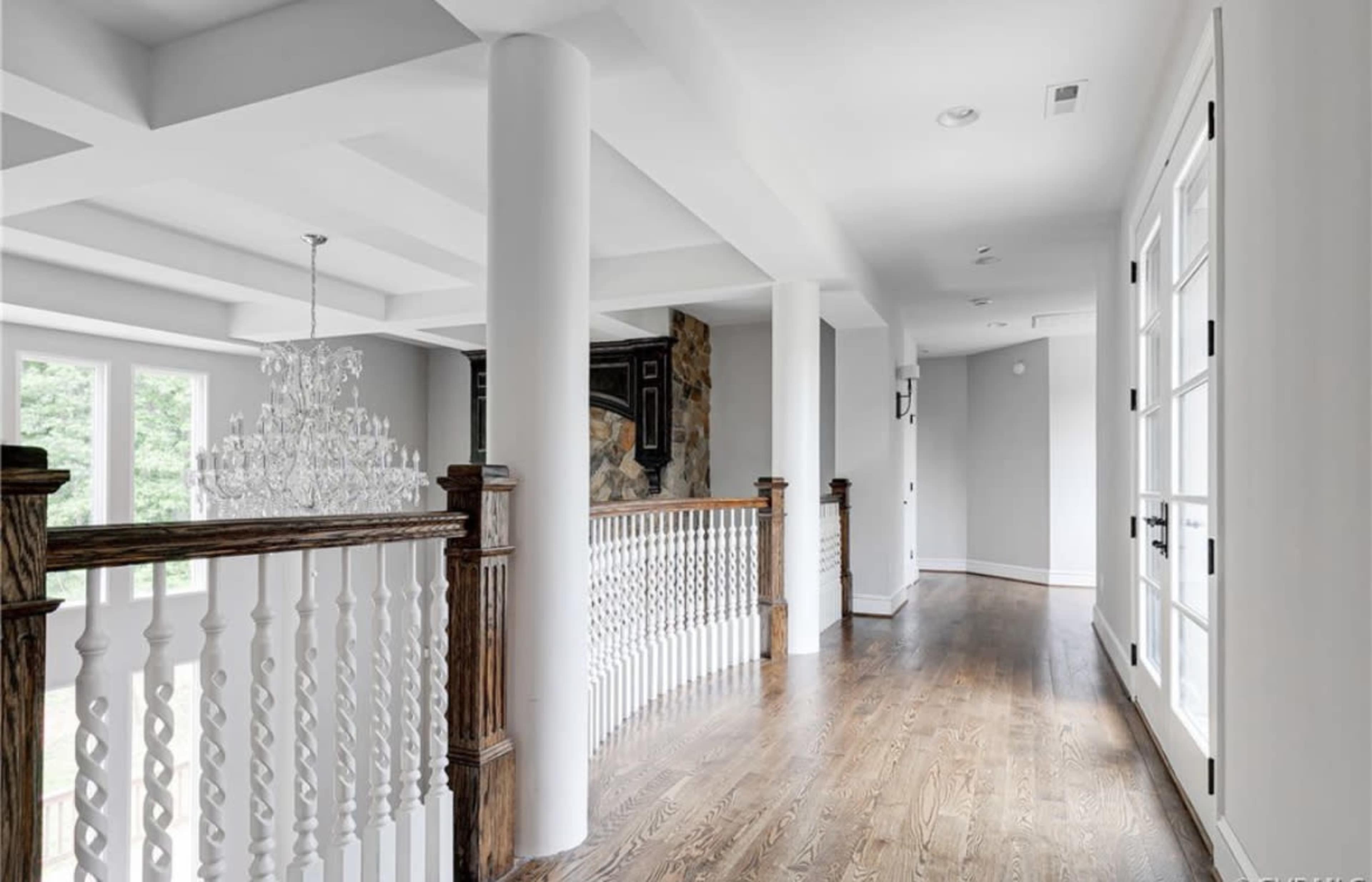 A spacious hallway with wooden flooring, white walls, and a decorative railing, illuminated by natural light coming through large windows.